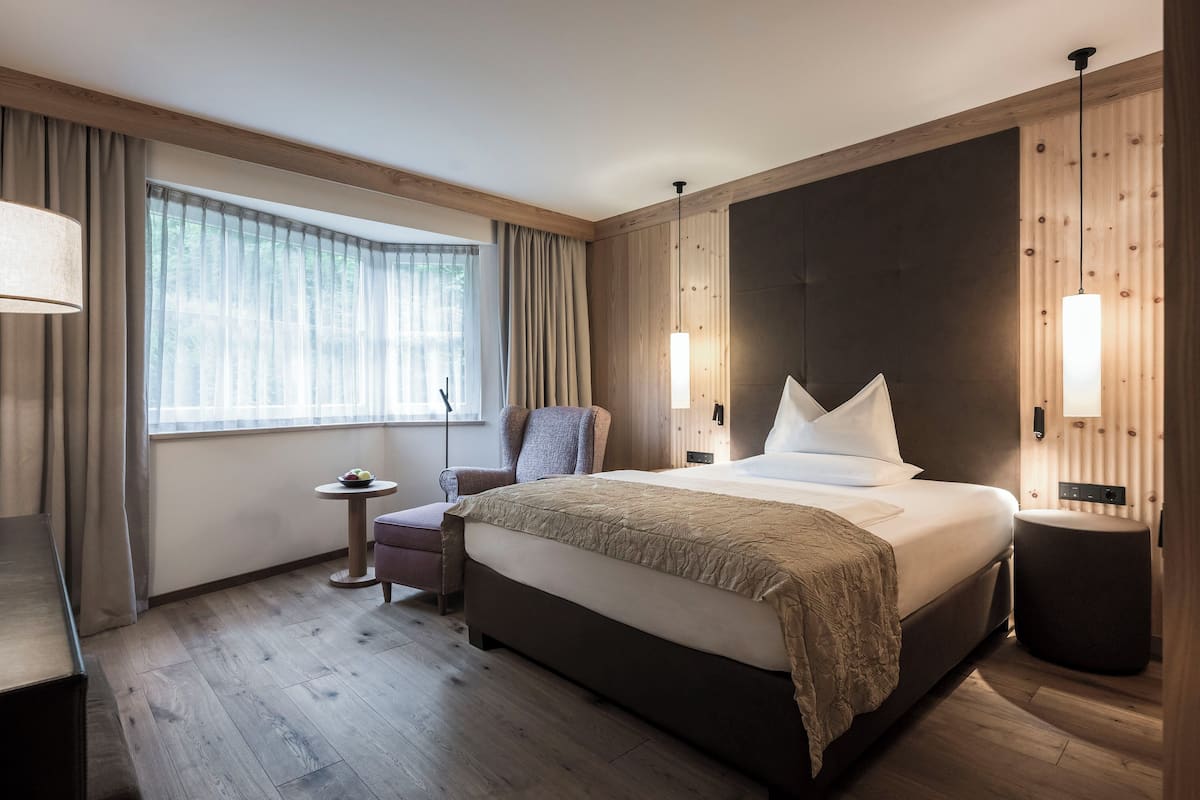 A modern hotel room with a large bed, a small round table, a chair, and a large window with curtains.