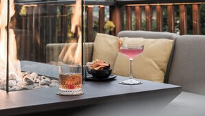 A table with a glass of pink cocktail, a glass of whiskey, and a bowl of snacks, set against a backdrop of a fire and mountains. - ADLER Dolomiti (Ortisei)