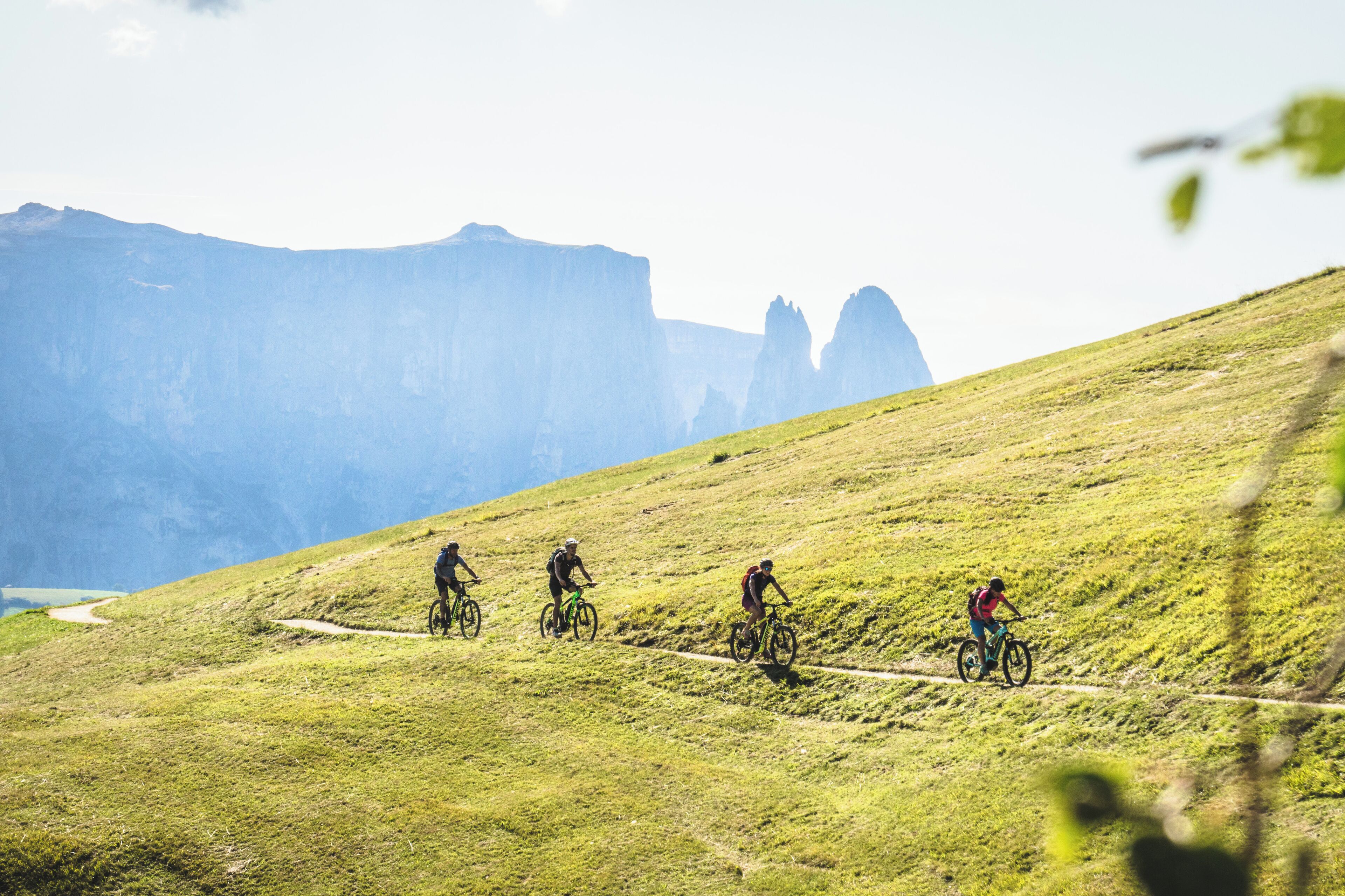 A group of cyclists on a grassy mountain trail with a clear sky and distant rocky formations.