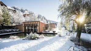 A modern wooden building with large windows, situated by a snow-covered landscape and a body of water. - ADLER Dolomiti (Ortisei)