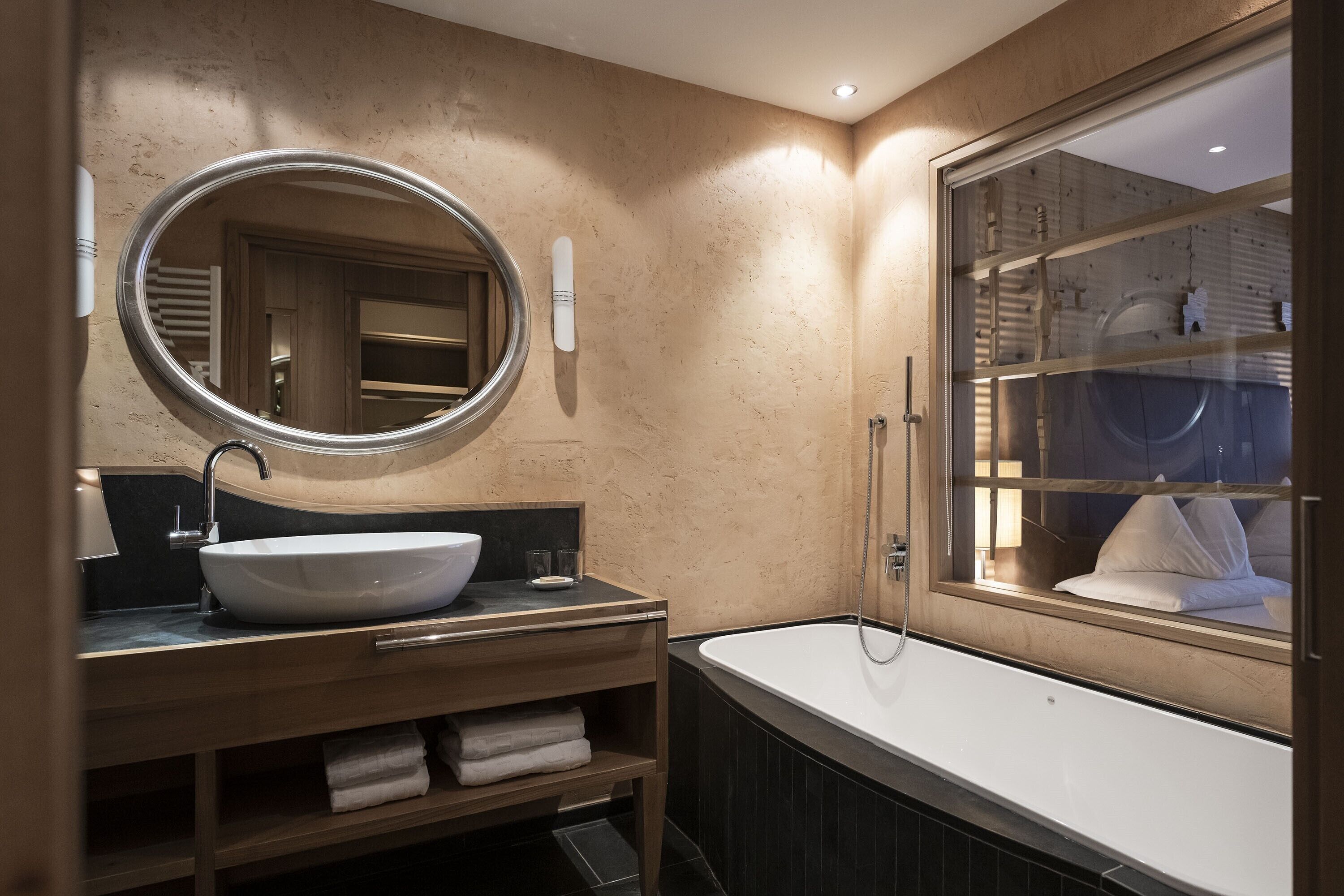Un baño moderno con un espejo ovalado grande, un lavamanos independiente, una bañera y una ventana con vistas a la habitación.