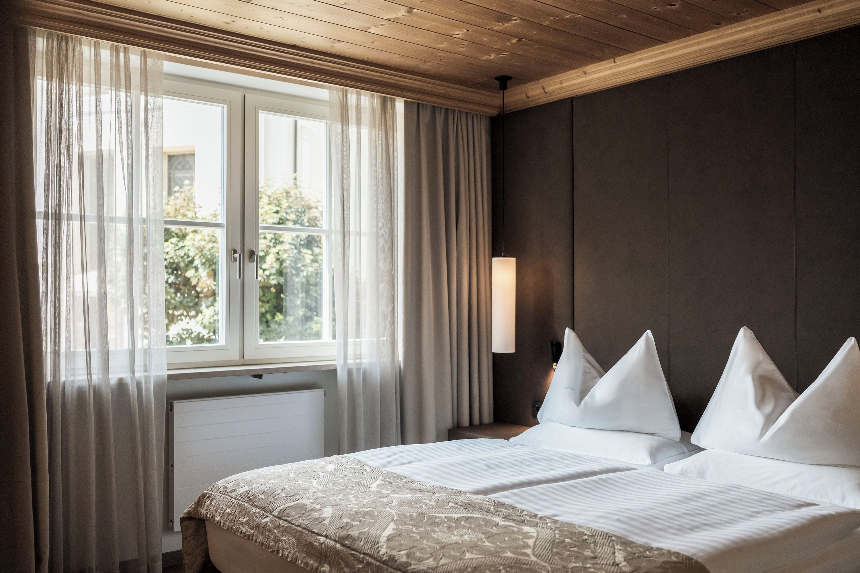 A hotel room with a large bed, a window with sheer curtains, a wall-mounted lamp, and a radiator.