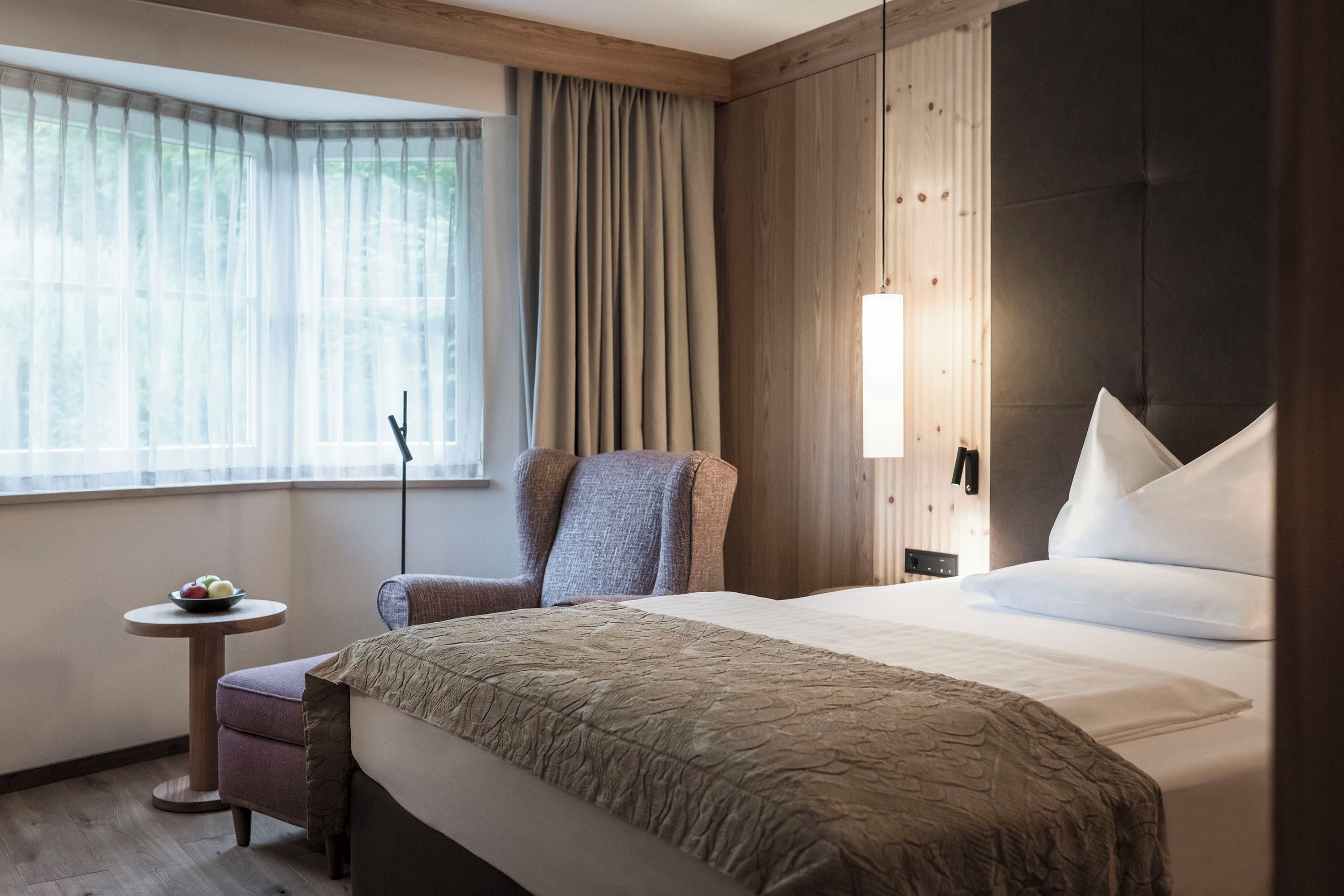 Ein Hotelzimmer mit einem großen Bett, einem Sessel, einem kleinen Tisch mit Früchten und einem Fenster mit Vorhängen.