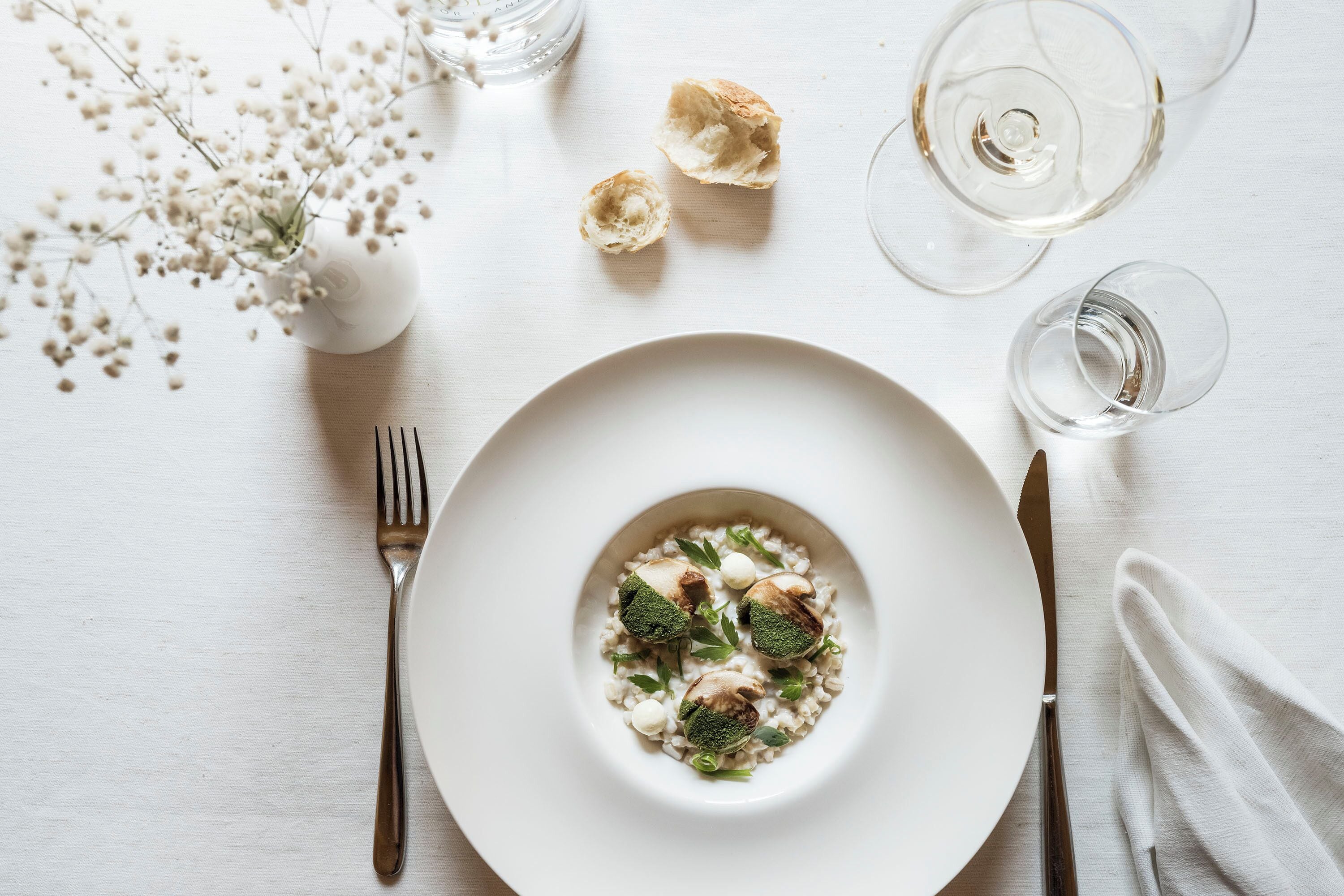 A white plate with a dish of food, a fork, a knife, and two wine glasses on a white table.