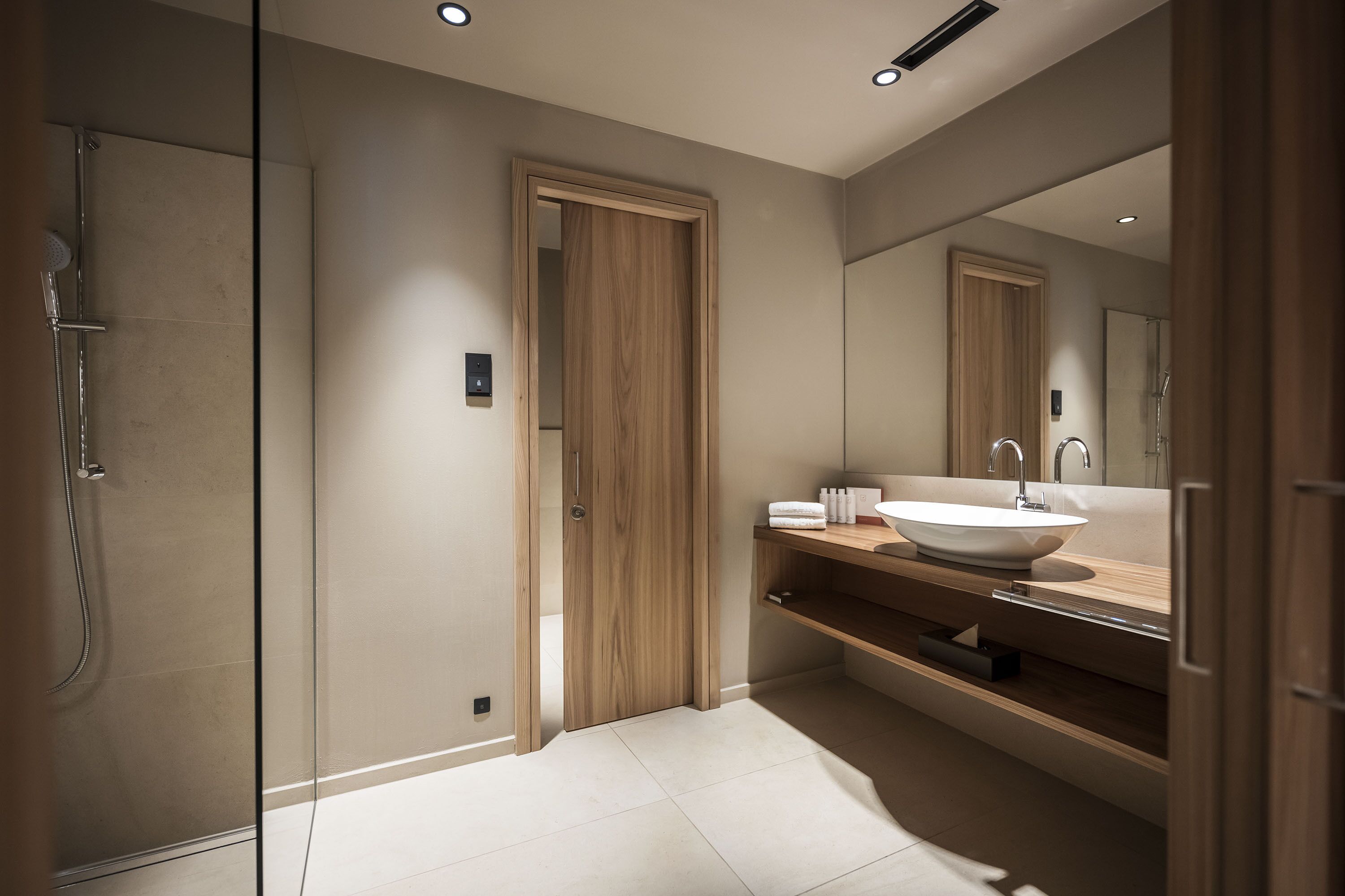 A modern bathroom with a glass shower enclosure, a wooden door, a large mirror, and a white sink with a wooden countertop.