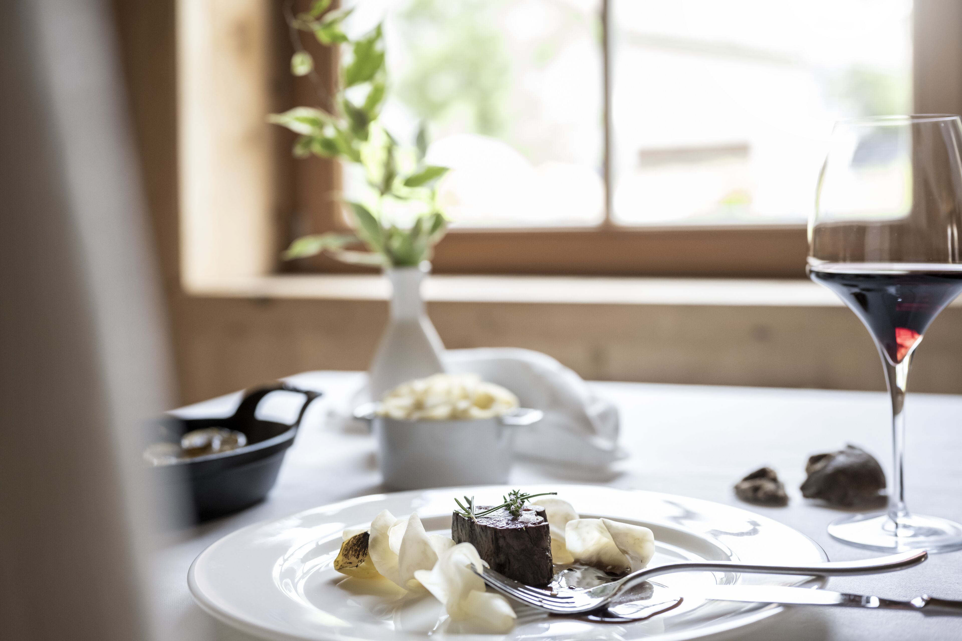 Une table à manger avec une assiette de nourriture, un verre de vin rouge et un vase avec une plante.