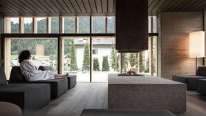 A modern interior with a person relaxing on a sofa, a fireplace, and large windows offering a view of the outdoors. - ADLER Dolomiti (Ortisei)