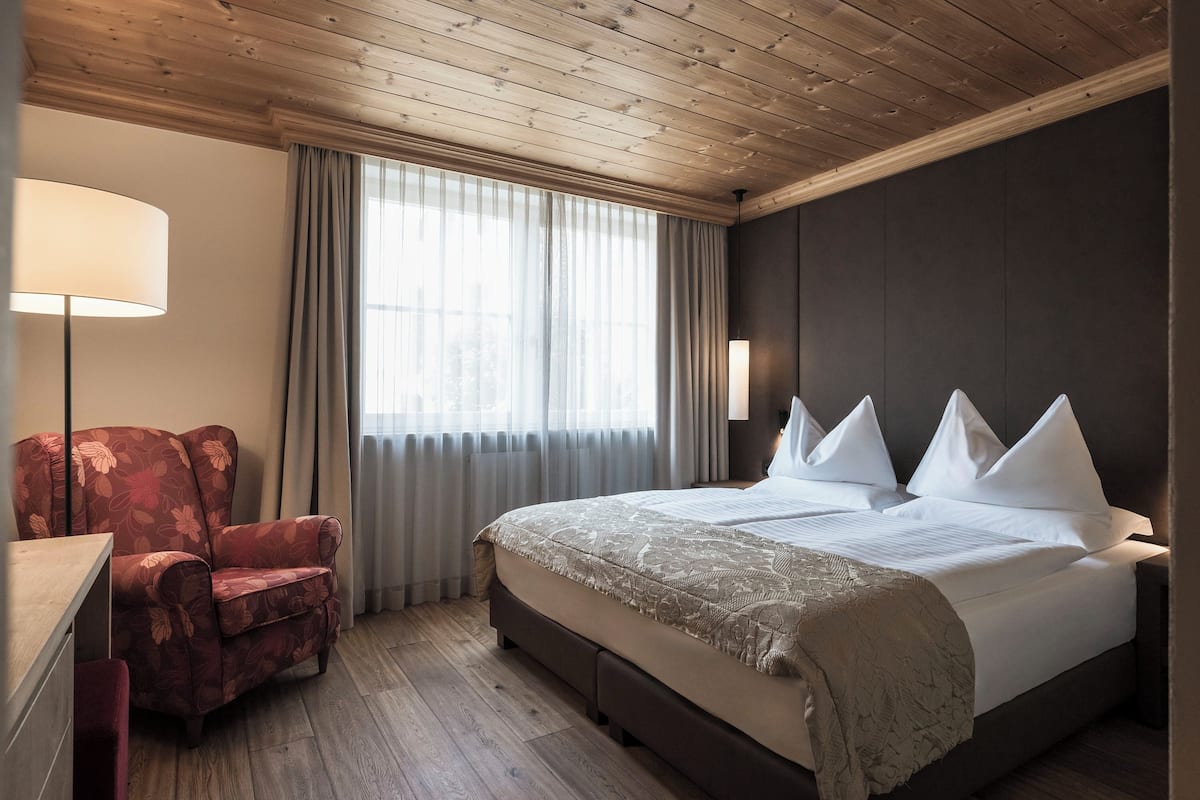 A hotel room with a wooden ceiling, a large bed with white pillows, a red armchair, and a desk with a lamp.