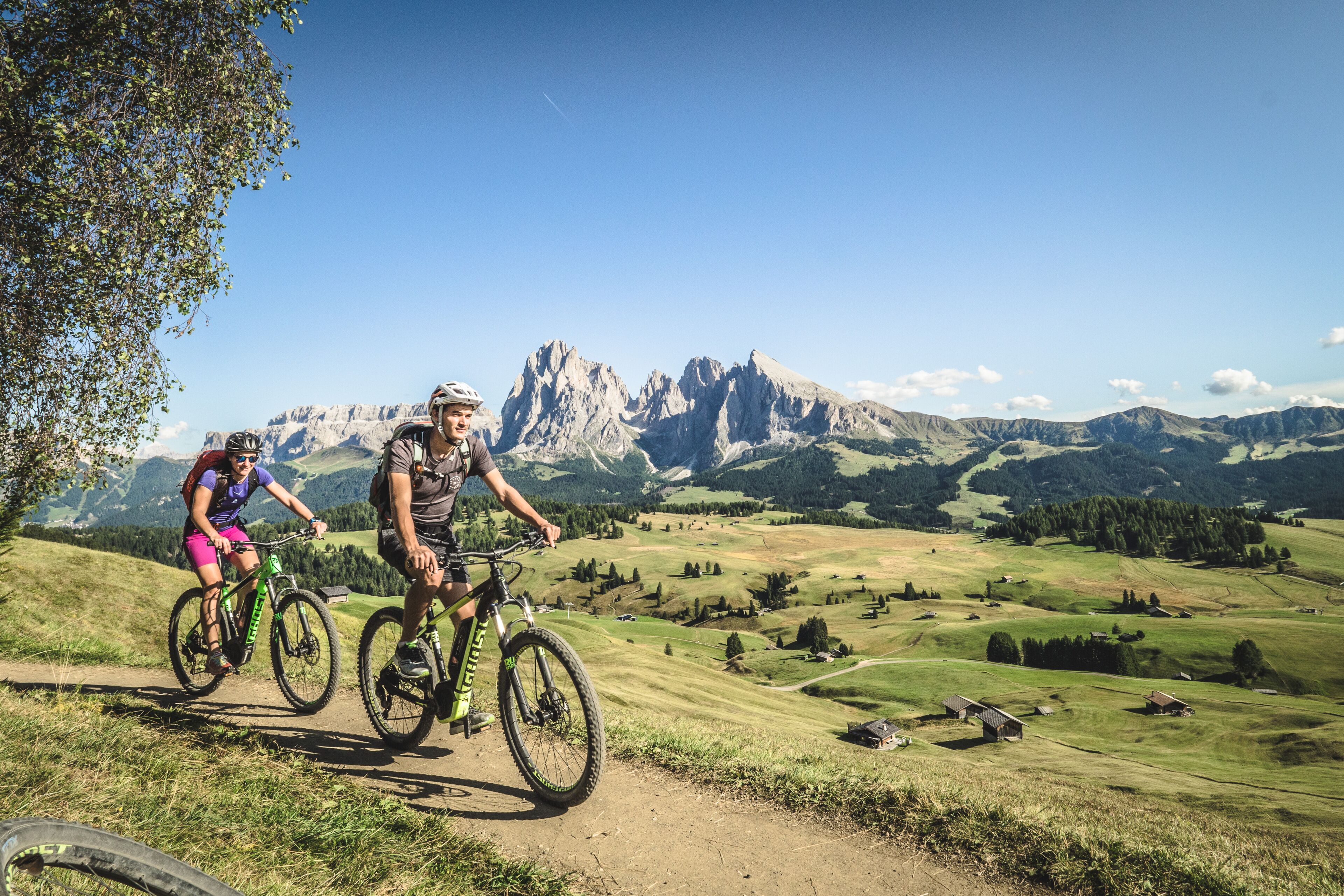 Two cyclists on a dirt path with a mountainous landscape in the background.