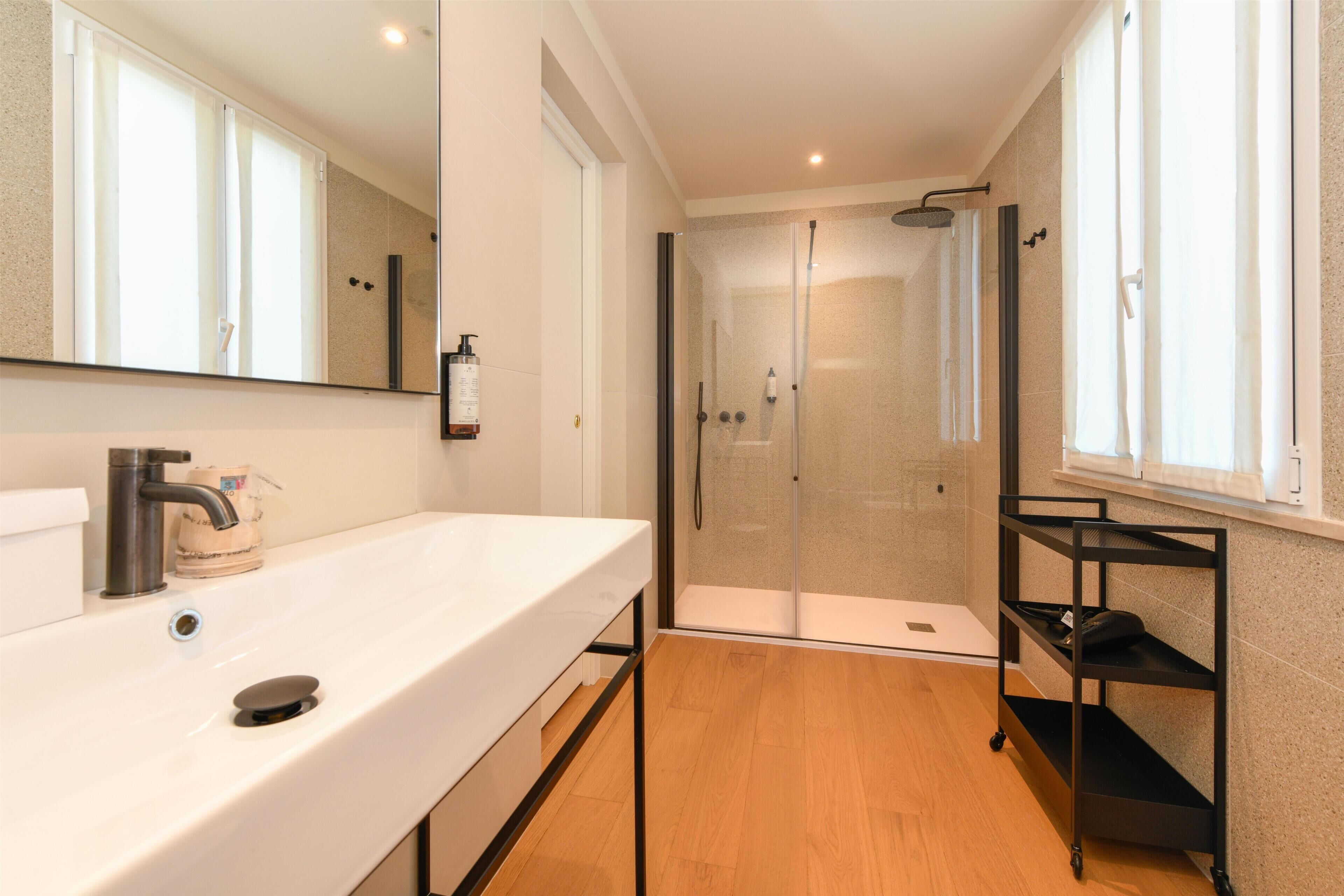 A modern bathroom with a white sink, a glass shower enclosure, and a black shelf unit.
