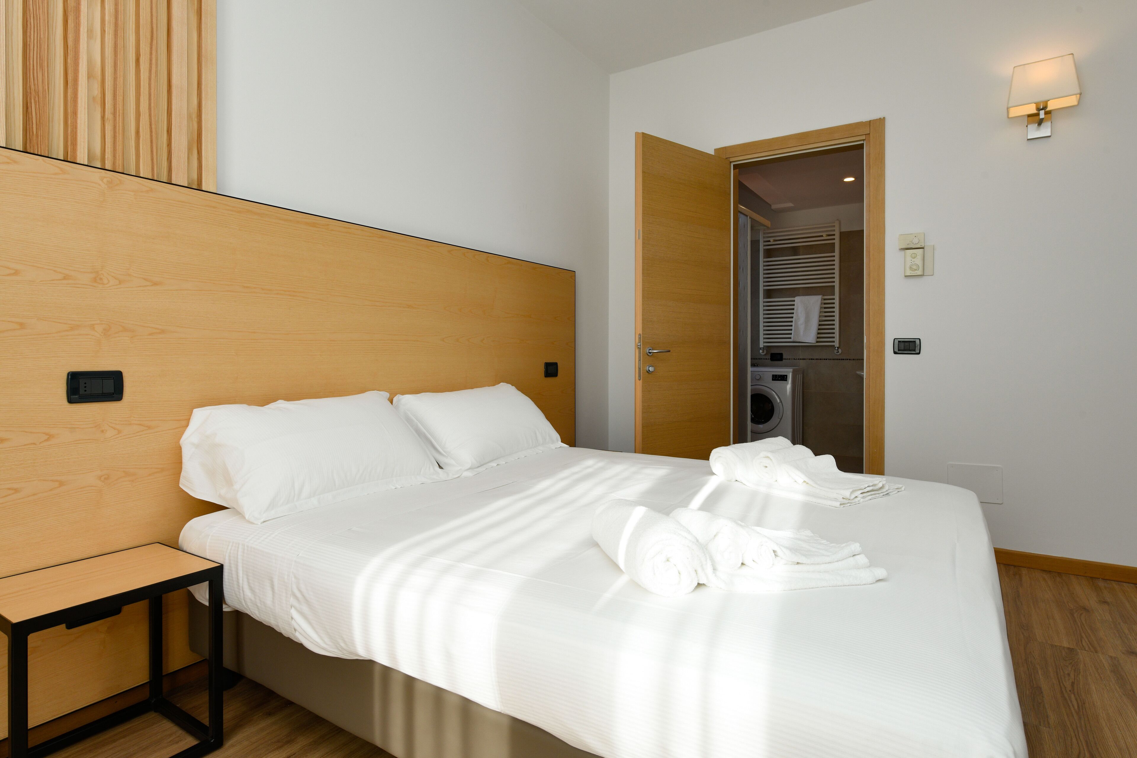 A hotel room with a wooden headboard, a bed with white linens, a small bedside table, and a visible bathroom with a towel rack.
