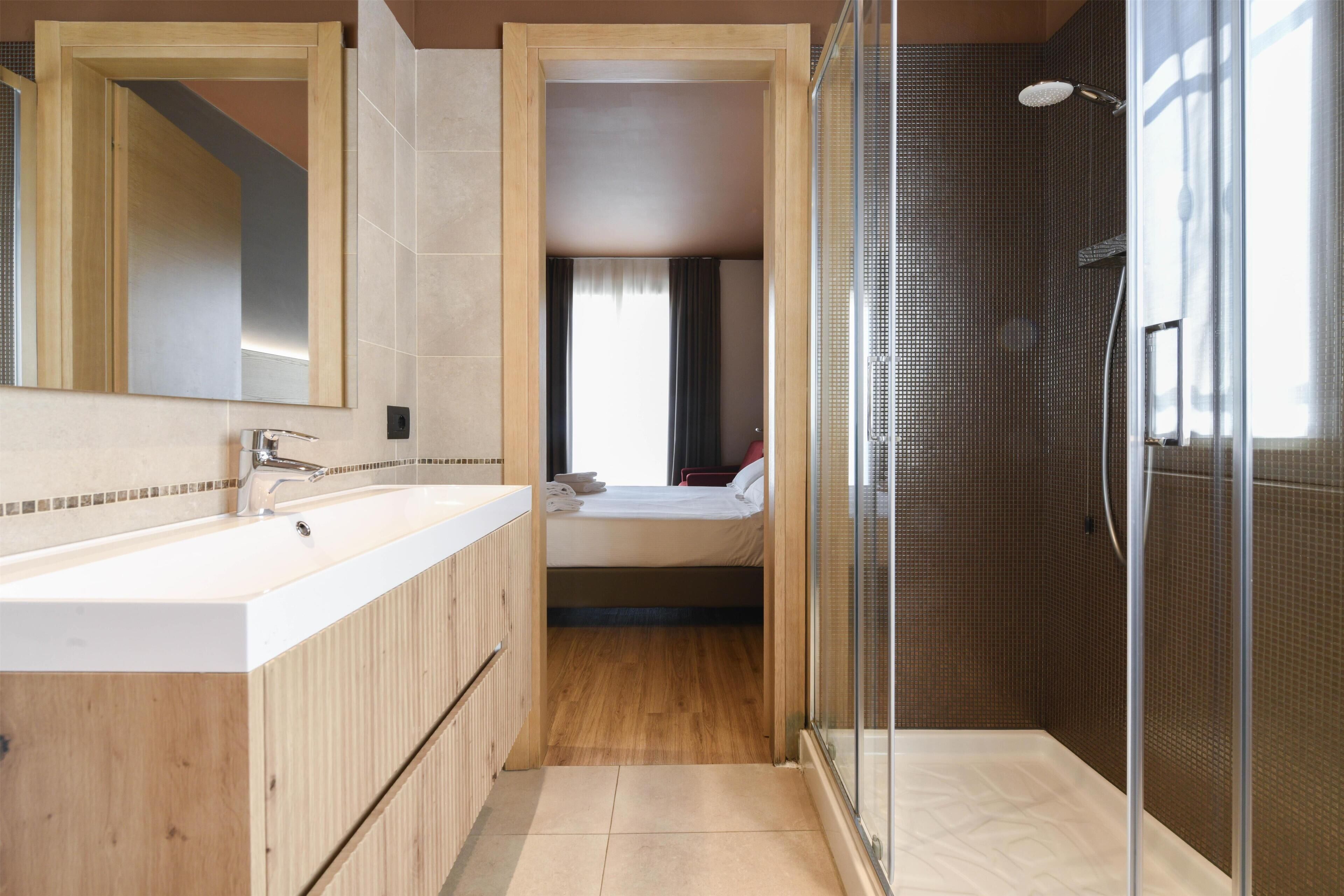 A modern bathroom with a large mirror, a shower enclosure, and a separate bedroom visible through an open door.