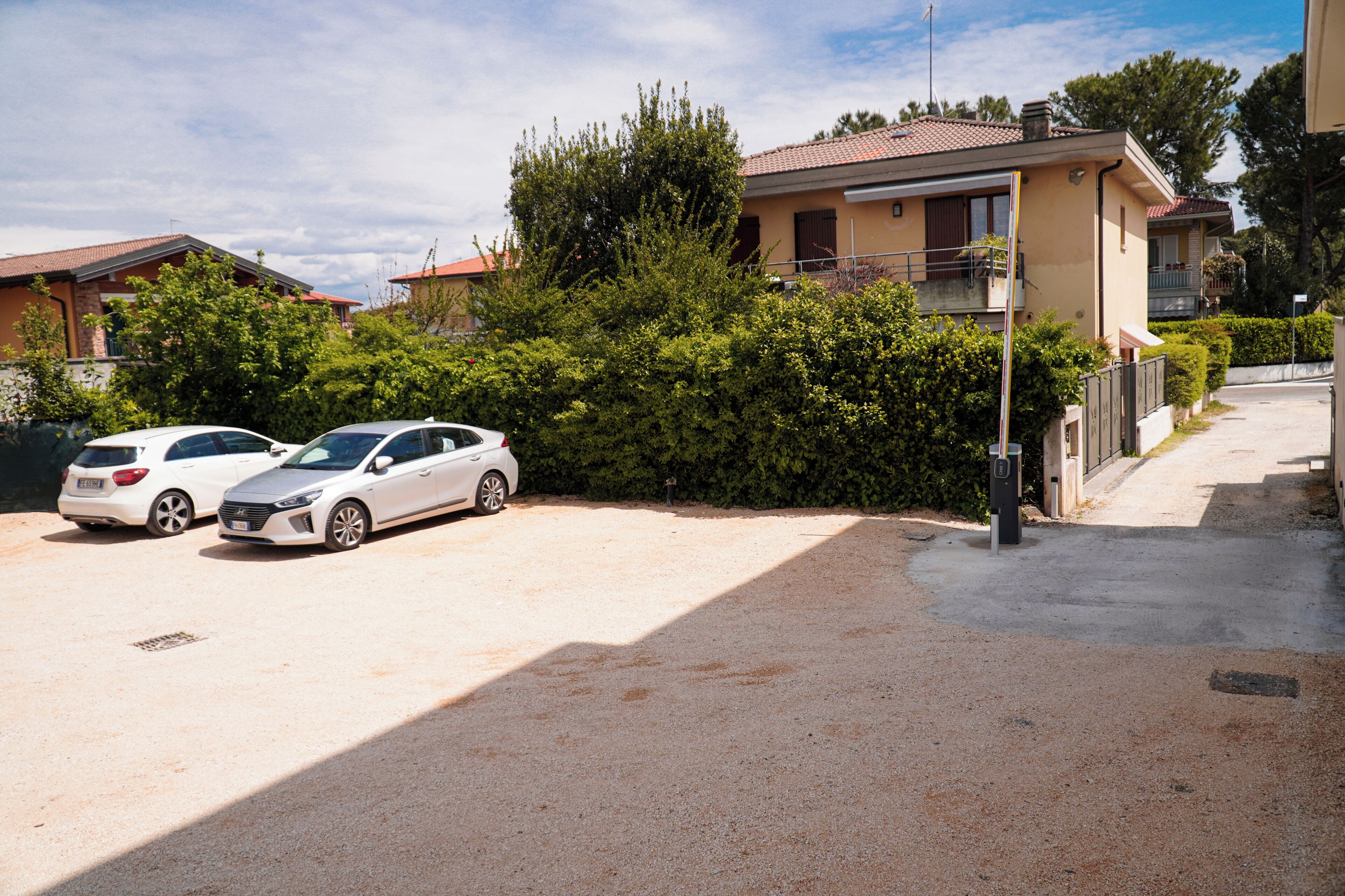 A residential area with two white cars parked in a driveway, surrounded by greenery and houses with red-tiled roofs.