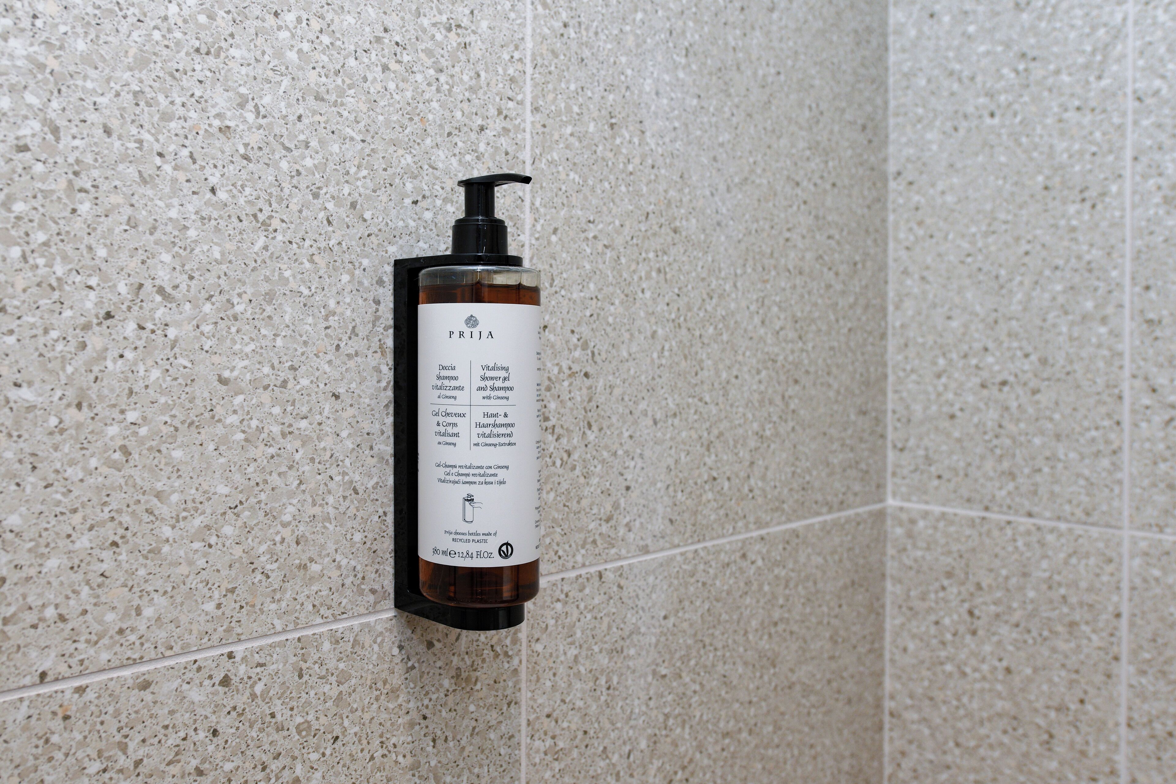 Une bouteille de shampoing et de gel douche de la marque PRJA, fixée sur un mur recouvert de carreaux à motifs.
