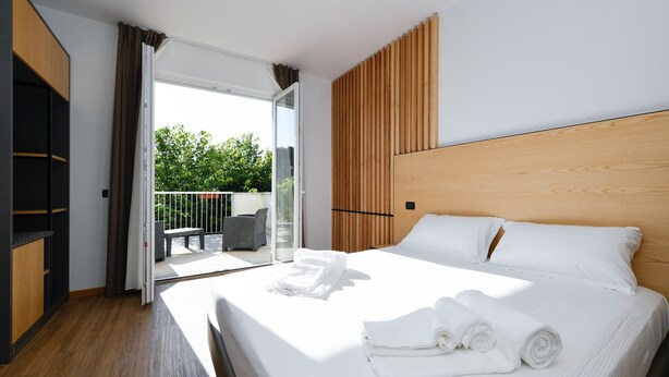 Quarto de hotel moderno com cama grande, cabeceira de madeira e varanda com mobiliário para exterior.