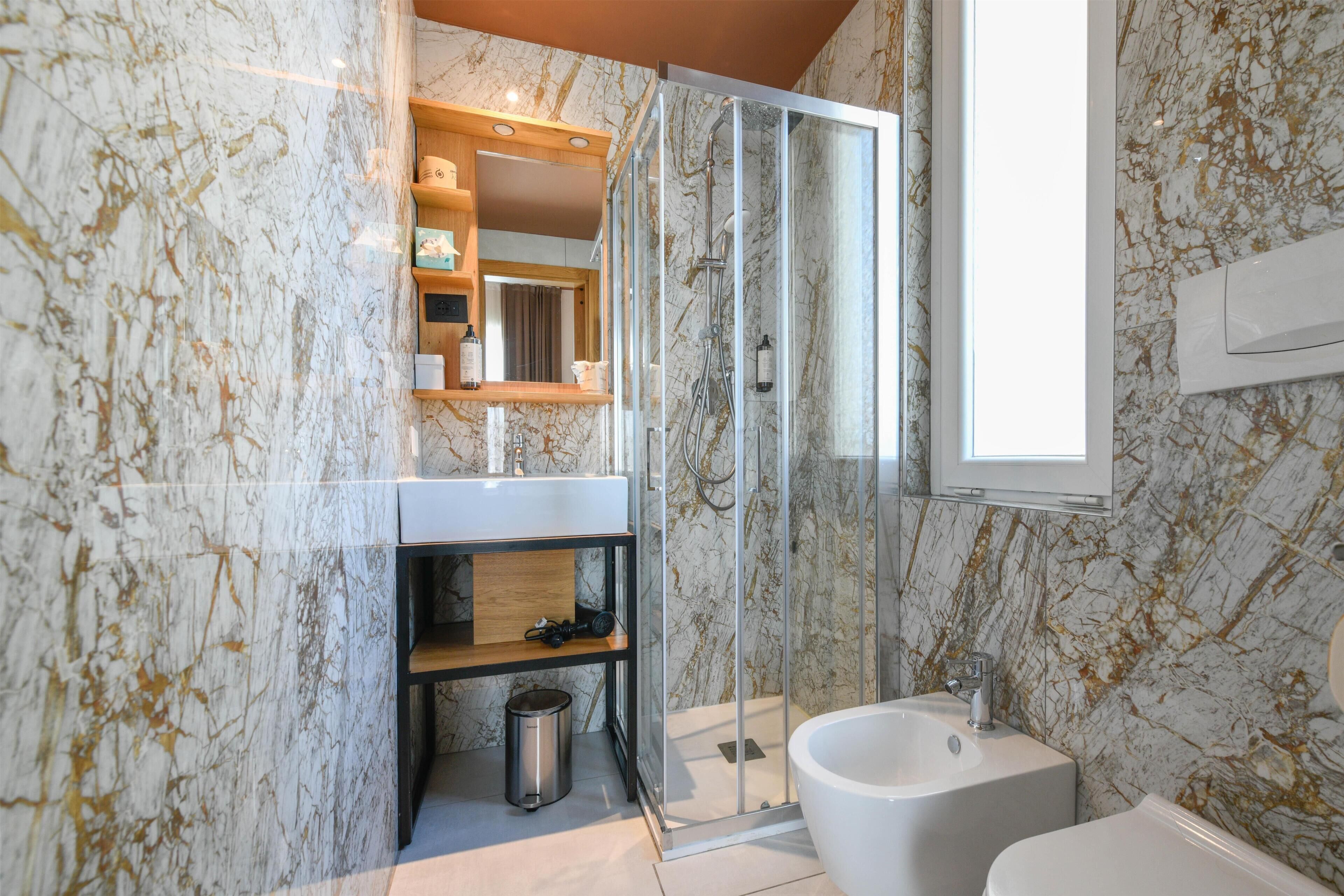 A bathroom with a marble wall, a glass shower enclosure, a sink, and a bidet.