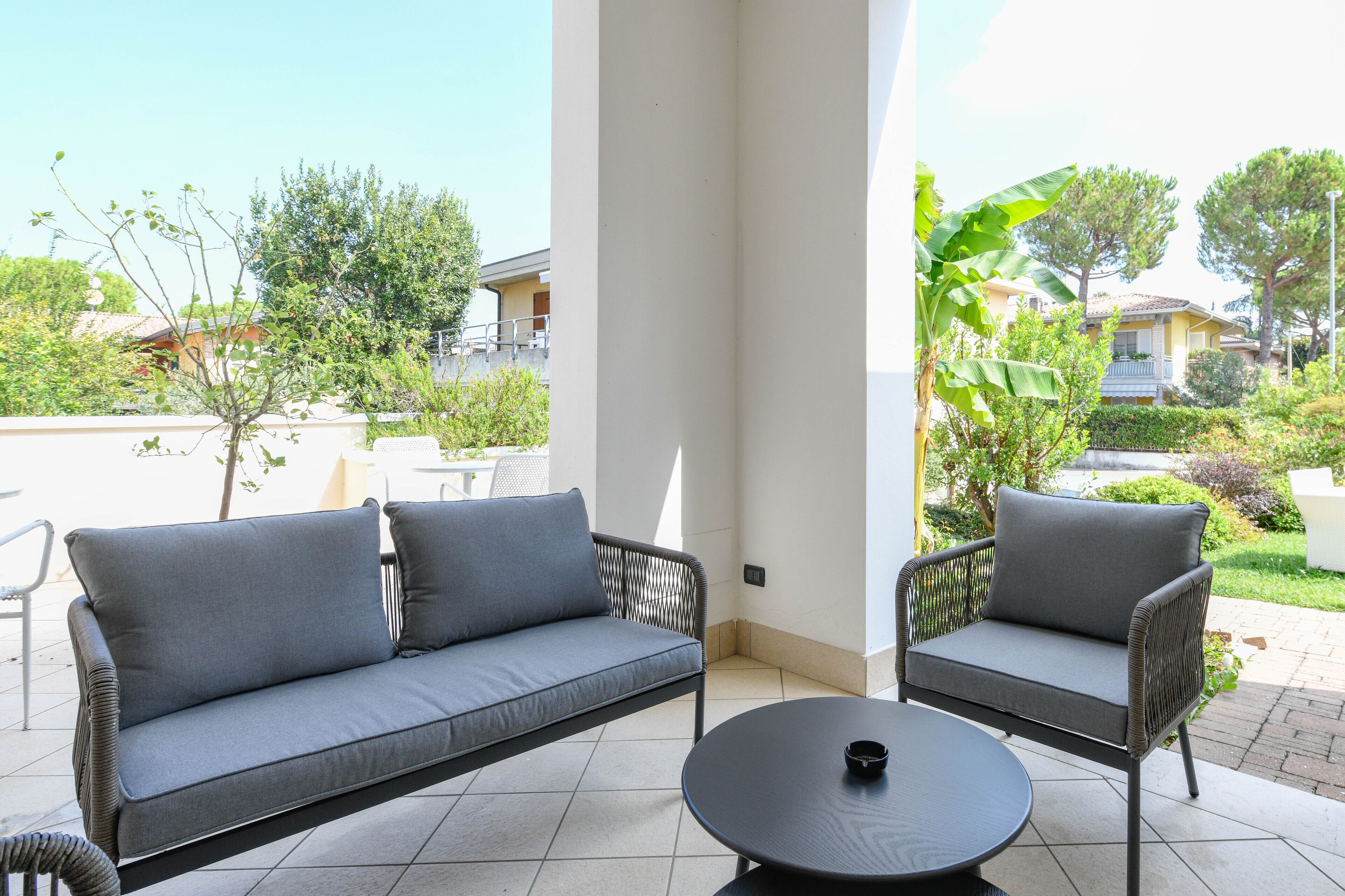 A patio area with a grey sofa and armchair, a round table, and a view of a residential neighborhood.