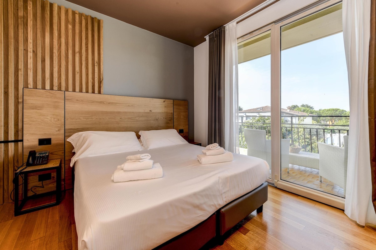 A hotel room with a large bed, a wooden headboard, a bedside table with a phone, and a balcony with a view.