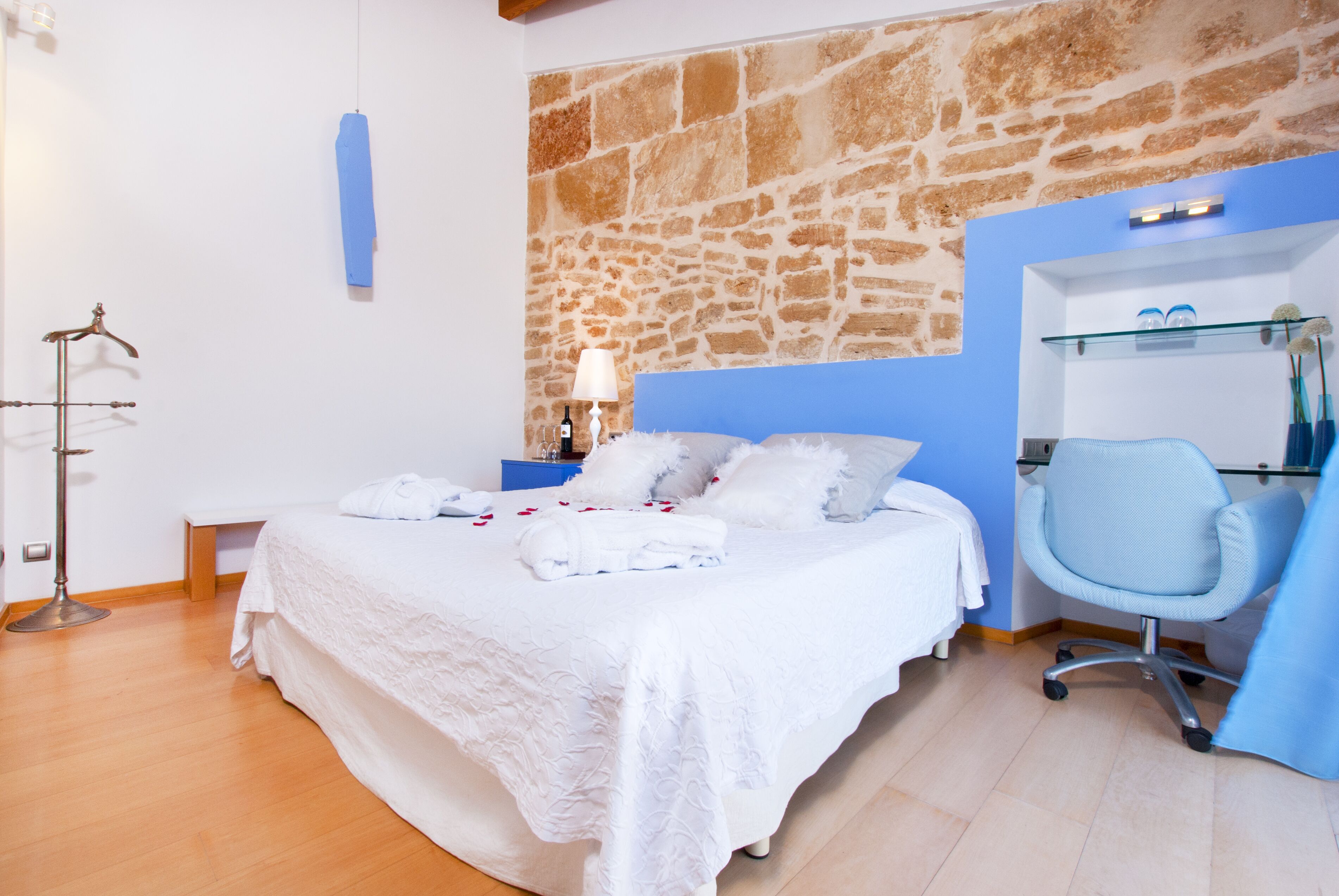 Un dormitorio con pared de piedra, cama con ropa de cama blanca, un escritorio con silla y un perchero.