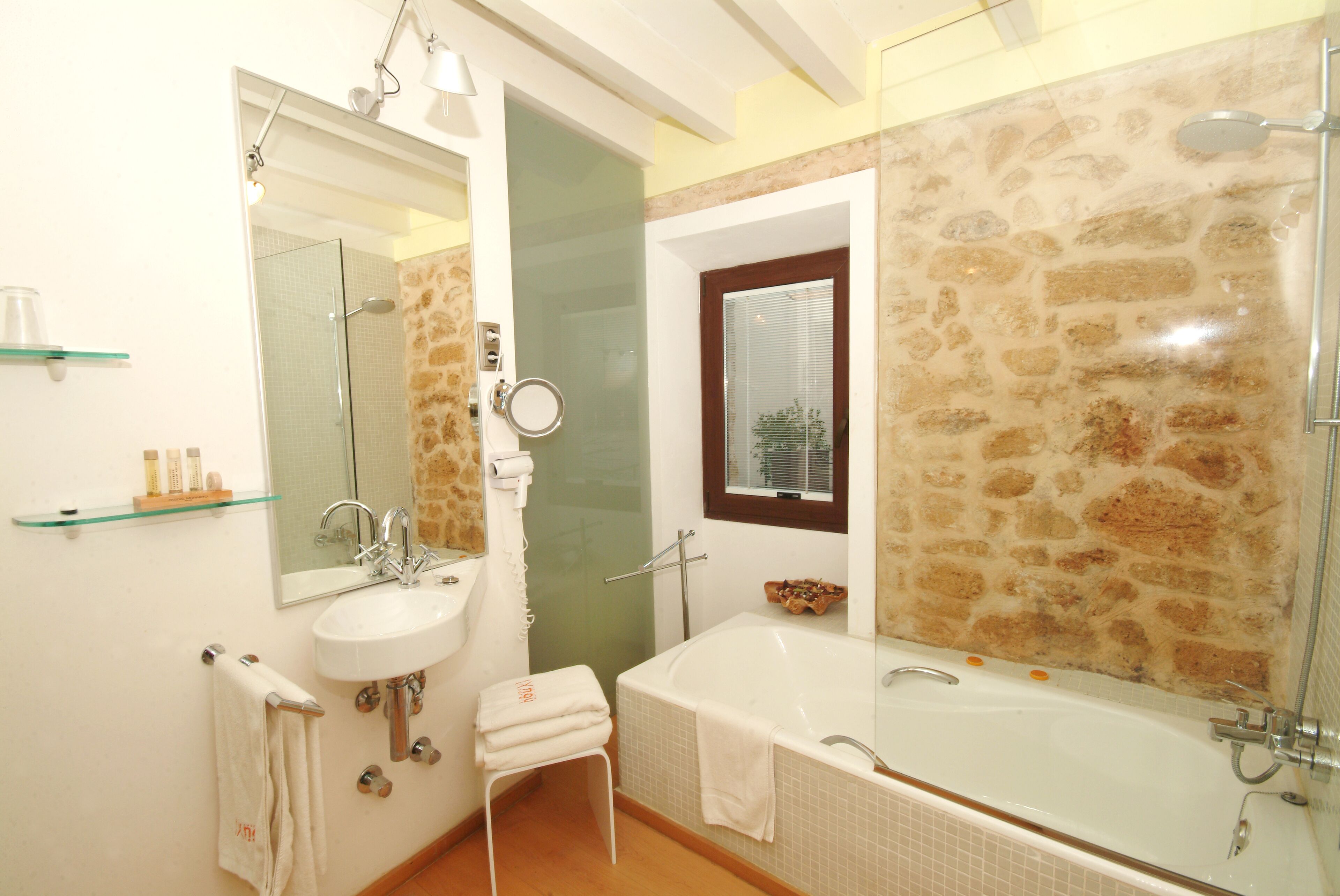Un baño con un lavamanos blanco, cabina de ducha de vidrio, pared de piedra y una ventana con persianas.