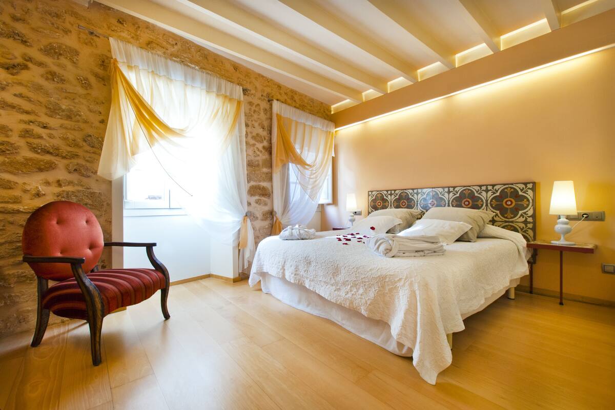 Une chambre avec un grand lit, un fauteuil rouge recouvert de tissu et un mur en pierre.