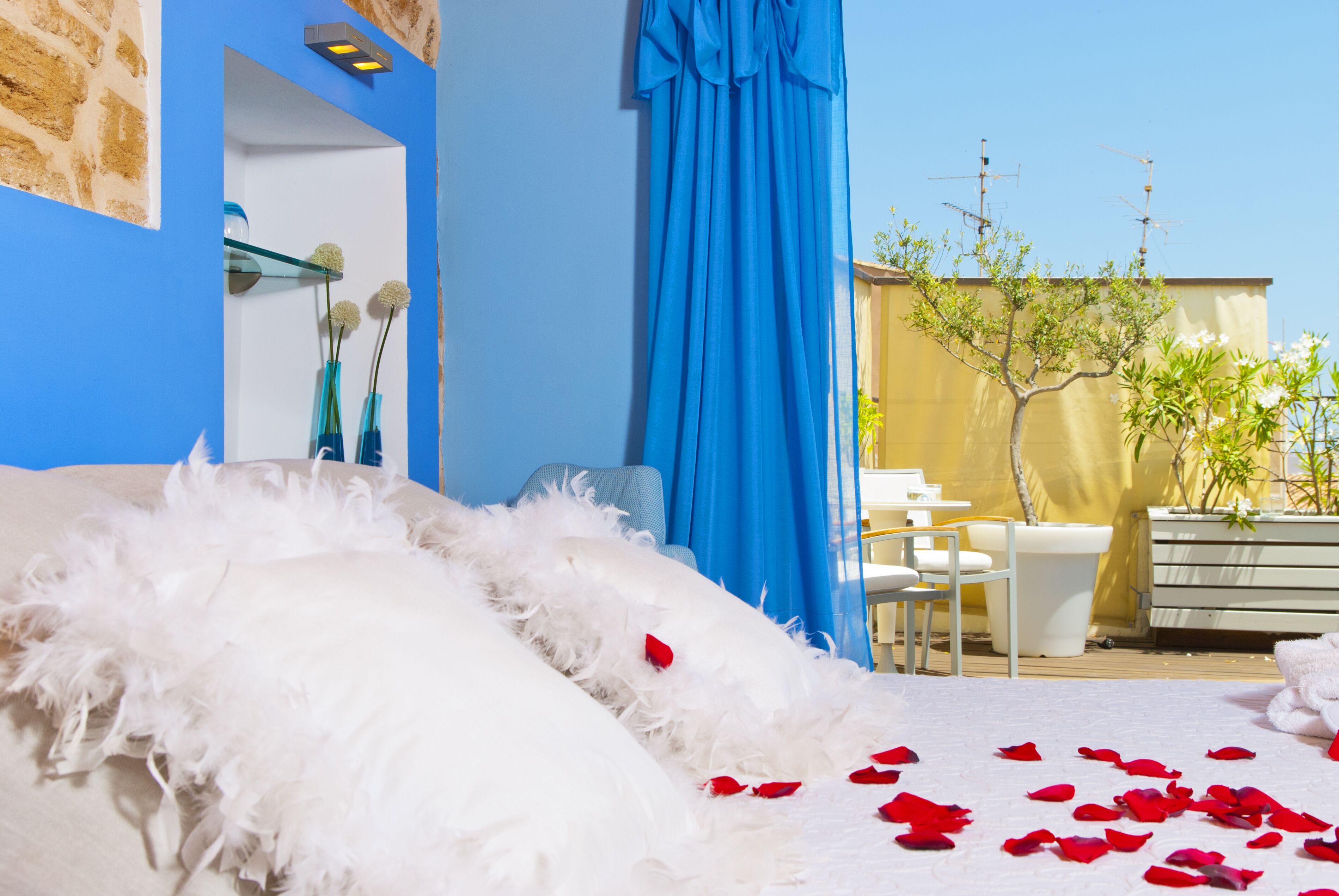 Un dormitorio con cama, plumas blancas, pétalos de rosa roja, pared azul, planta en maceta y ventana con cortinas.