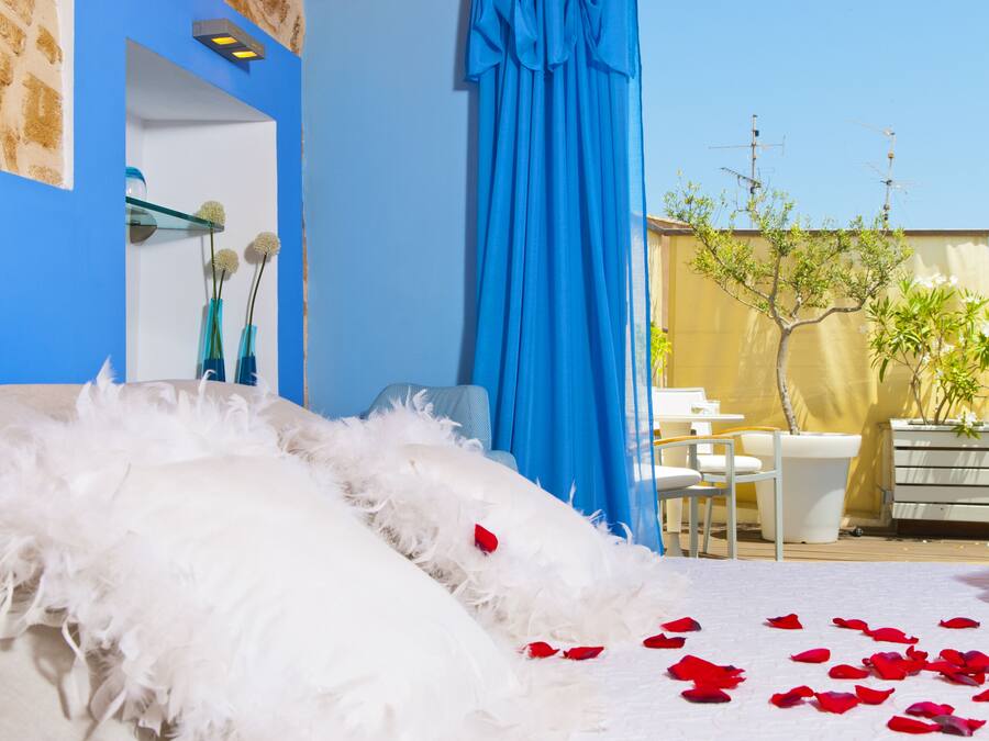 Un dormitorio con cama, plumas blancas, pétalos de rosa roja, pared azul, planta en maceta y ventana con cortinas.