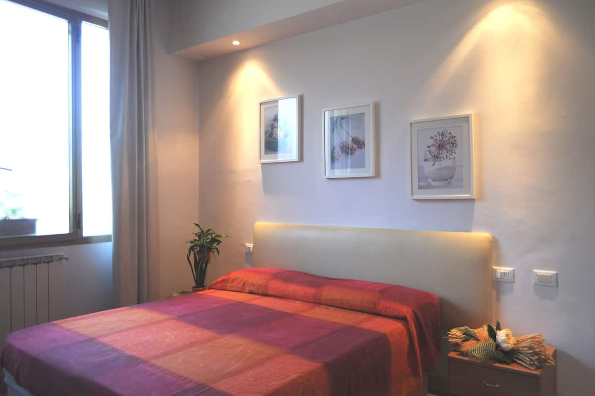 A bedroom with a bed, a nightstand, a potted plant, and two framed pictures on the wall.