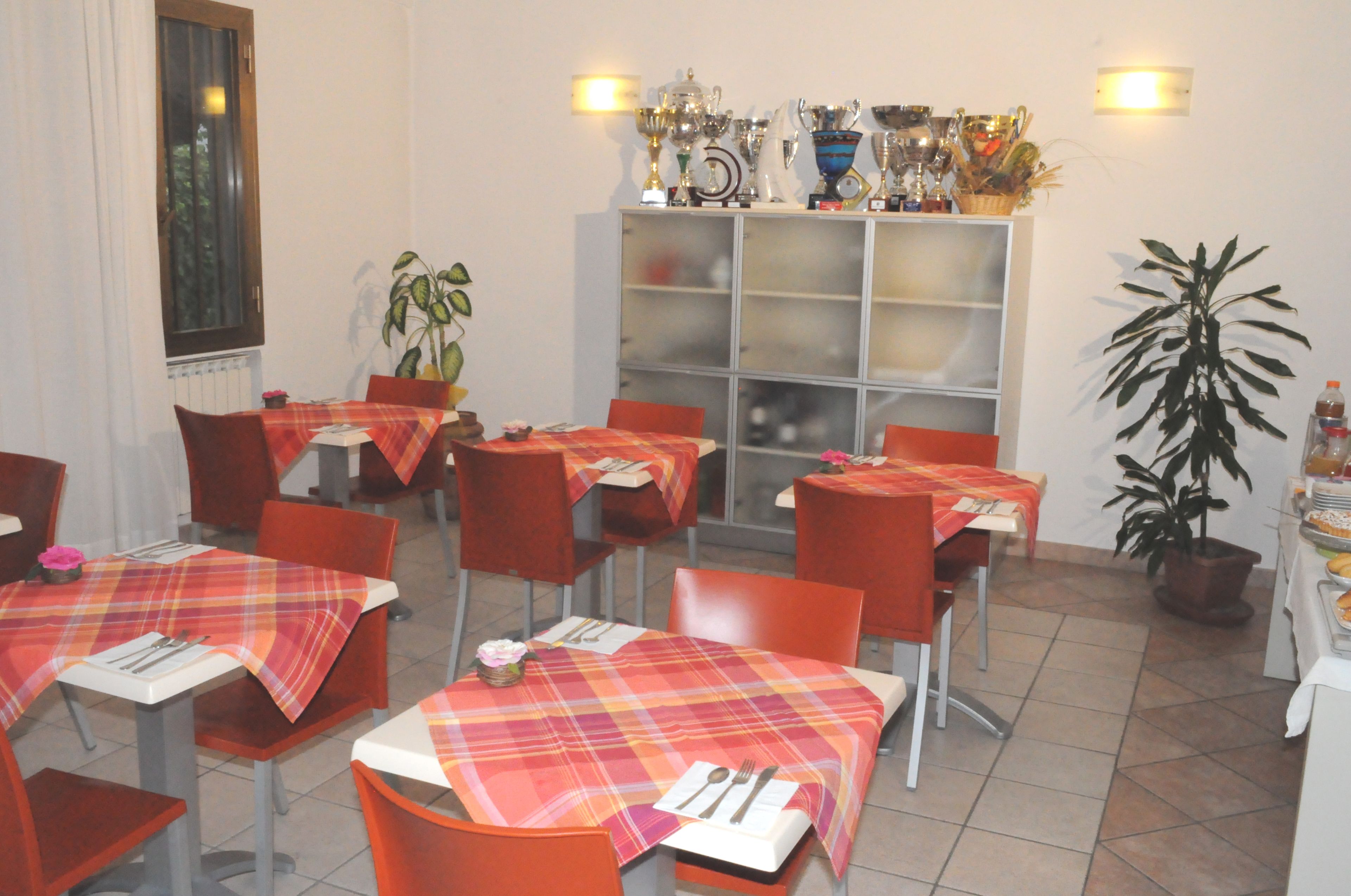 Een restaurant met meerdere tafels, bedekt met geruit tafelkleed, rode stoelen en een vitrine met trofeeën aan de muur.