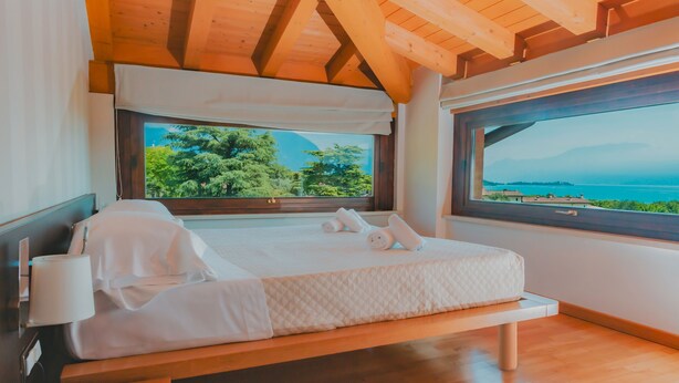 Ein Schlafzimmer mit Holzboden, einem großen Fenster mit Blick auf Bäume und ein Gewässer, einem Bett mit weißer Bettwäsche und einem Nachttisch mit Lampe und Telefon.