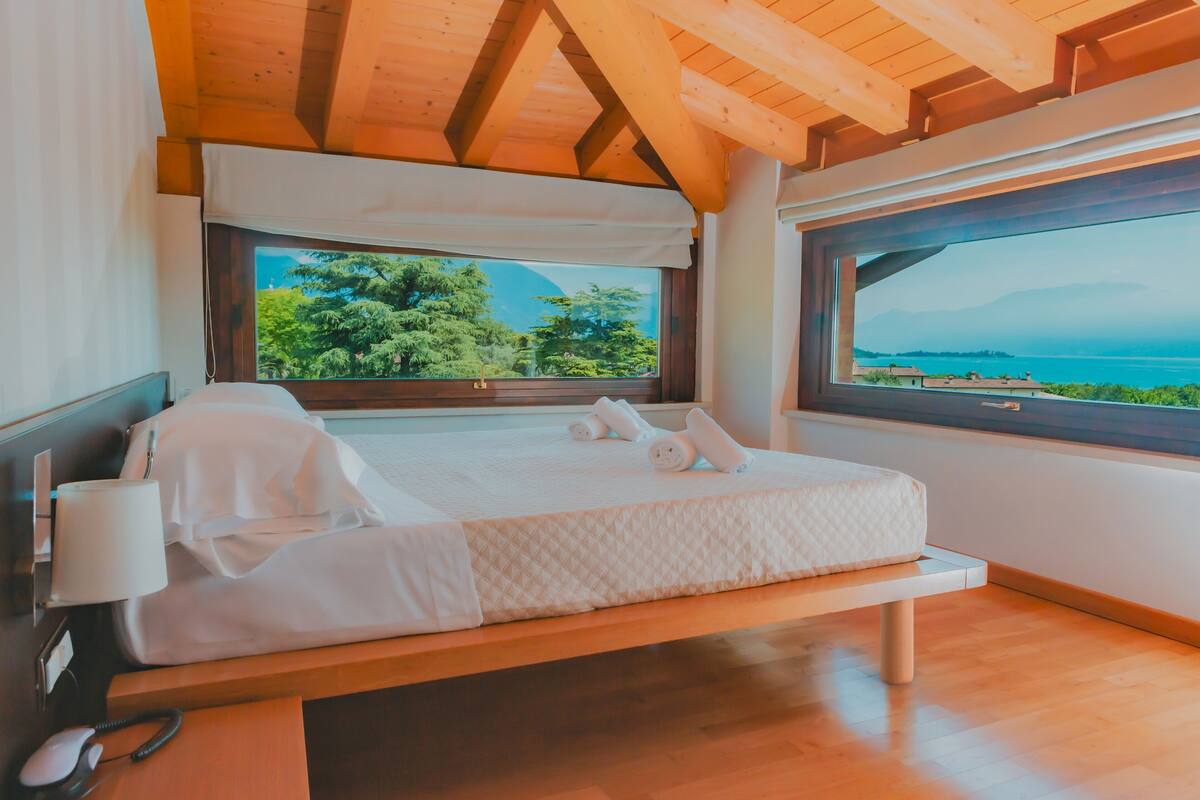 Un dormitorio con techo de madera, un ventanal con vistas a árboles y un cuerpo de agua, una cama con ropa de cama blanca y una mesita de noche con lámpara y teléfono.