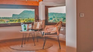 Ein Zimmer mit Holzboden, zwei Korbstühlen, einem kleinen runden Tisch und Blick auf eine grüne Landschaft durch große Fenster.