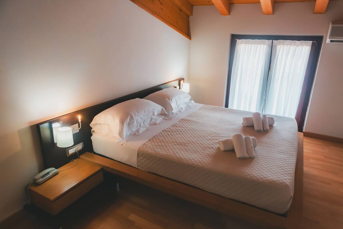 Una cama bien hecha con sábanas blancas y toallas enrolladas, una lámpara de noche y un teléfono sobre una mesita de madera.