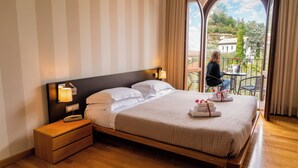 Ein ordentlich bezogenes Bett mit weißen Laken, ein Nachttisch mit Lampe und Blick auf einen Balkon mit Tisch und Stühlen.