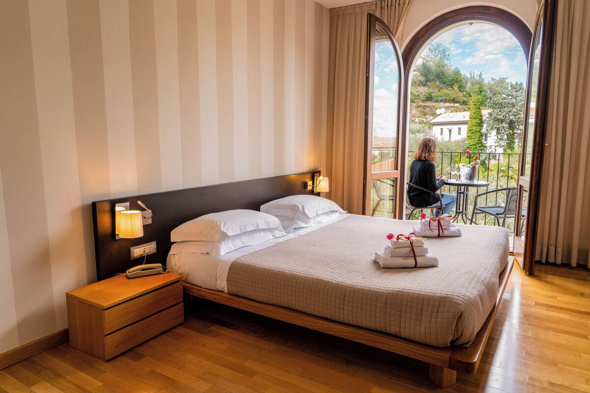Una cama bien hecha con ropa blanca, una mesita de noche con una lámpara y vistas a un balcón con mesa y sillas.