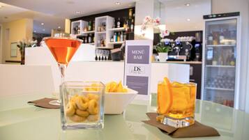 Una zona de bar con una copa de martini, un bol de aperitivos y una copa de bebida color ámbar sobre una mesa de vidrio.