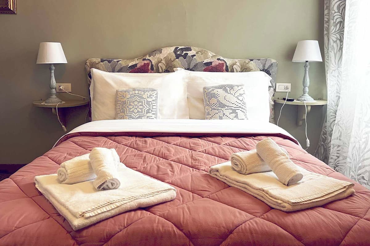 Quarto de hotel com cama, duas luminárias de cabeceira, mesa de cabeceira e janela com cortinas.