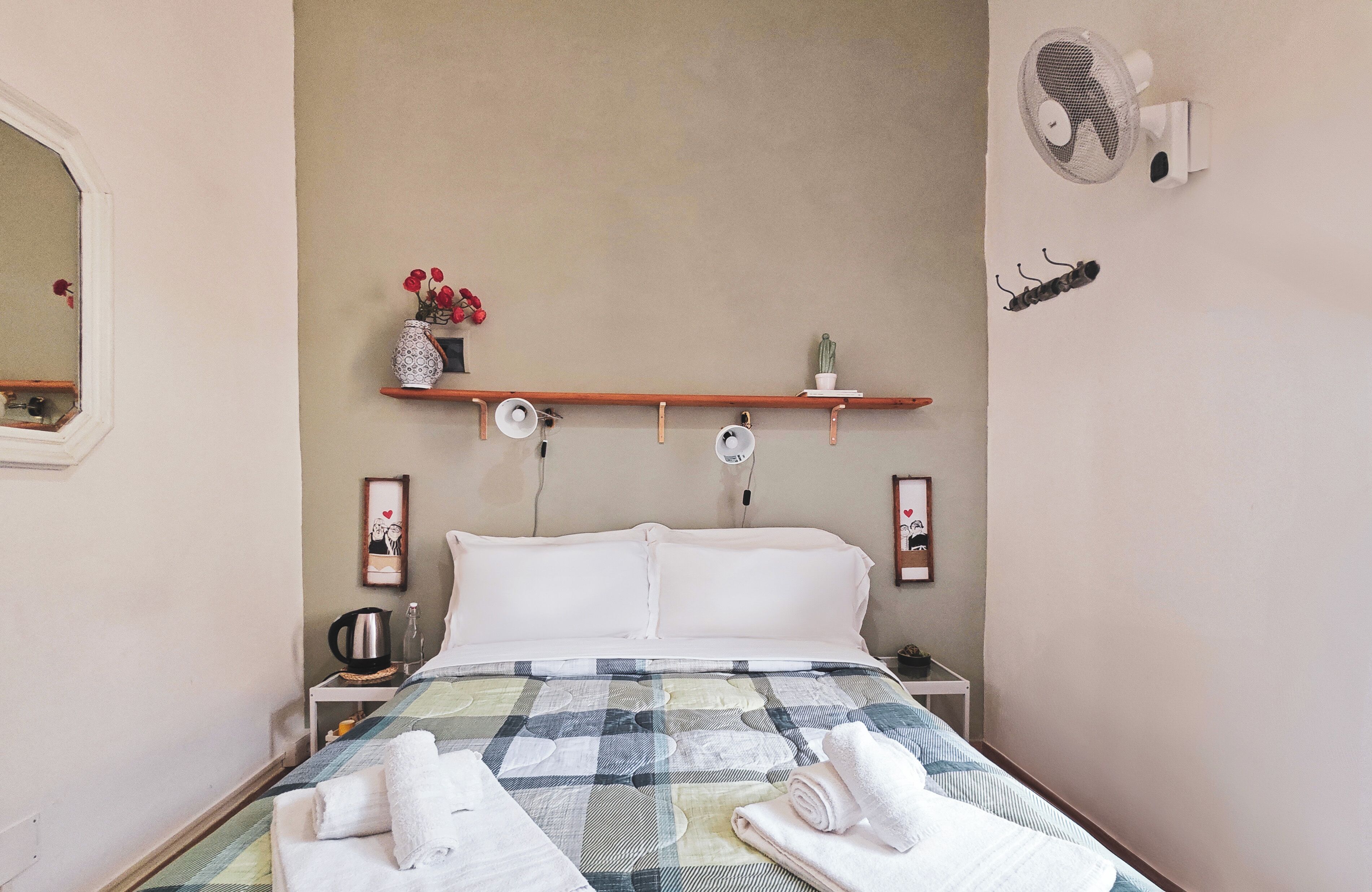 A neatly made bed with a checkered bedspread, two white pillows, and rolled towels on the side. There is a shelf above the bed holding a vase with flowers and a small cactus. A wall-mounted fan and a decorative item resembling a caterpillar are also visible.