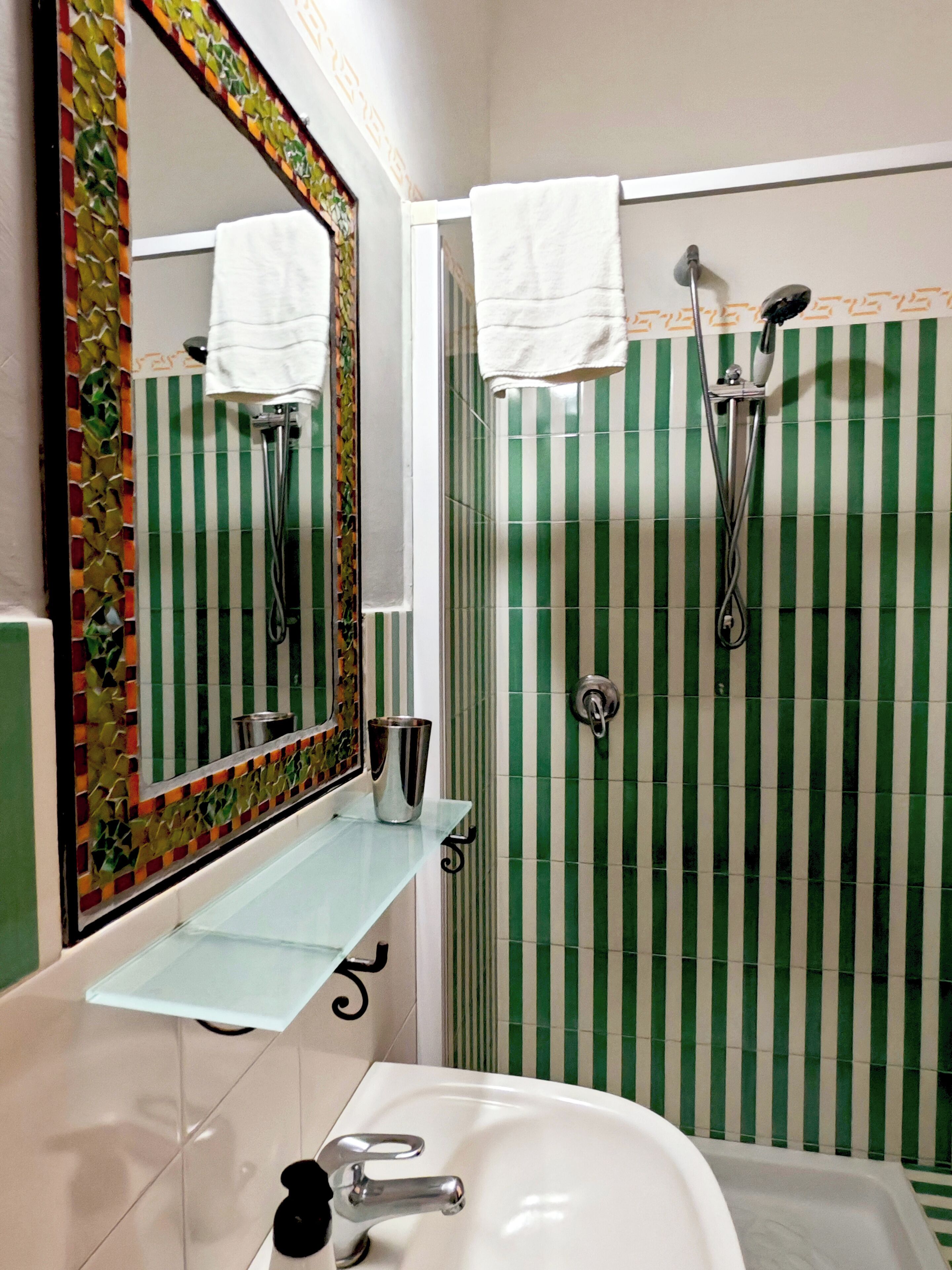 A bathroom with a glass shelf, a white sink, a mirror, and a shower with green-striped tiles.