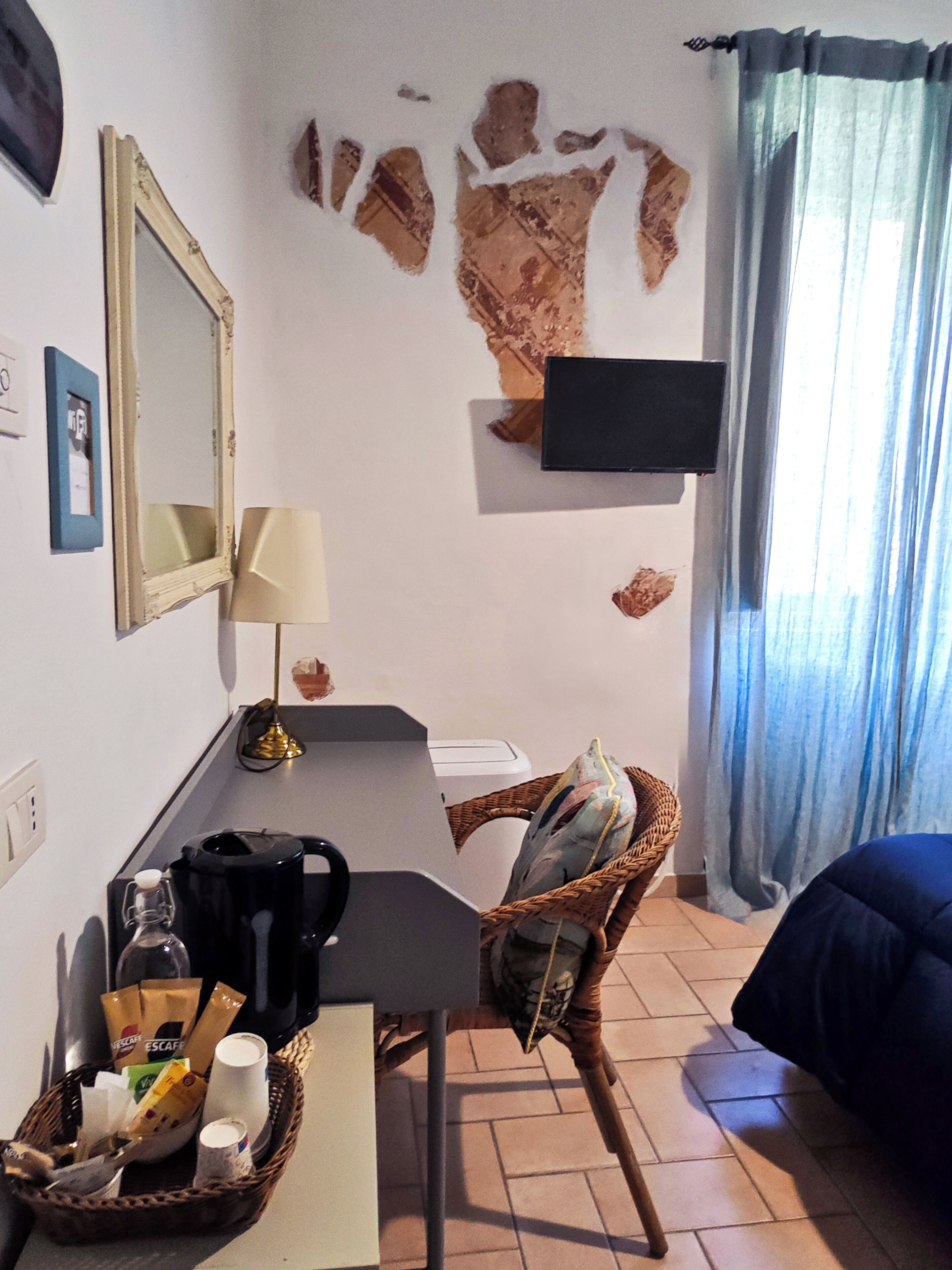 Ein Hotelzimmer mit Schreibtisch, Stuhl und Bett. Ein Fernseher ist an der Wand befestigt, eine Lampe vorhanden und ein Korb mit Pflegeprodukten.