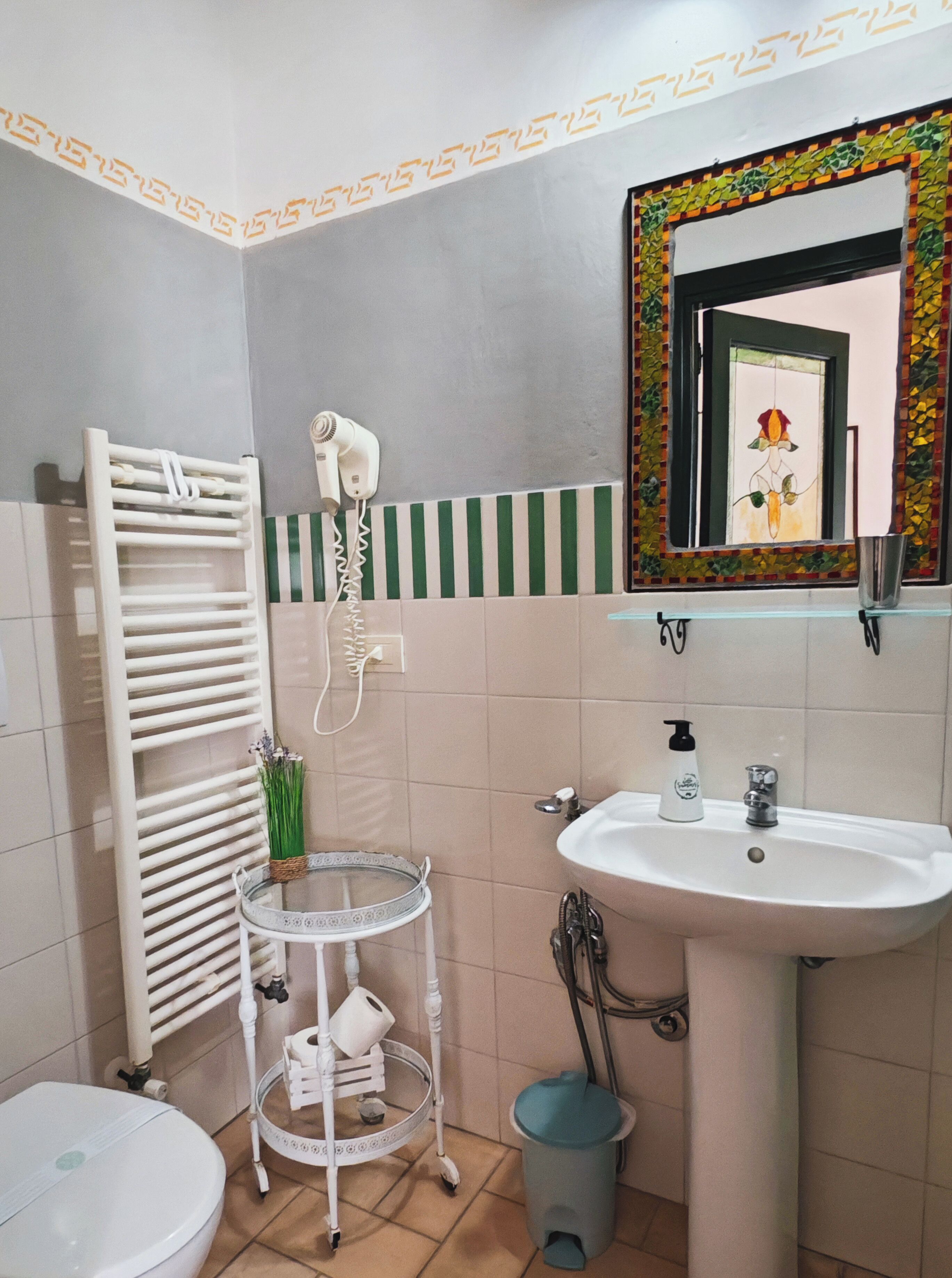 Un baño con un lavabo blanco, un espejo con un marco decorativo, un perchero para toallas, un secador de pelo y una mesita redonda con una cesta.