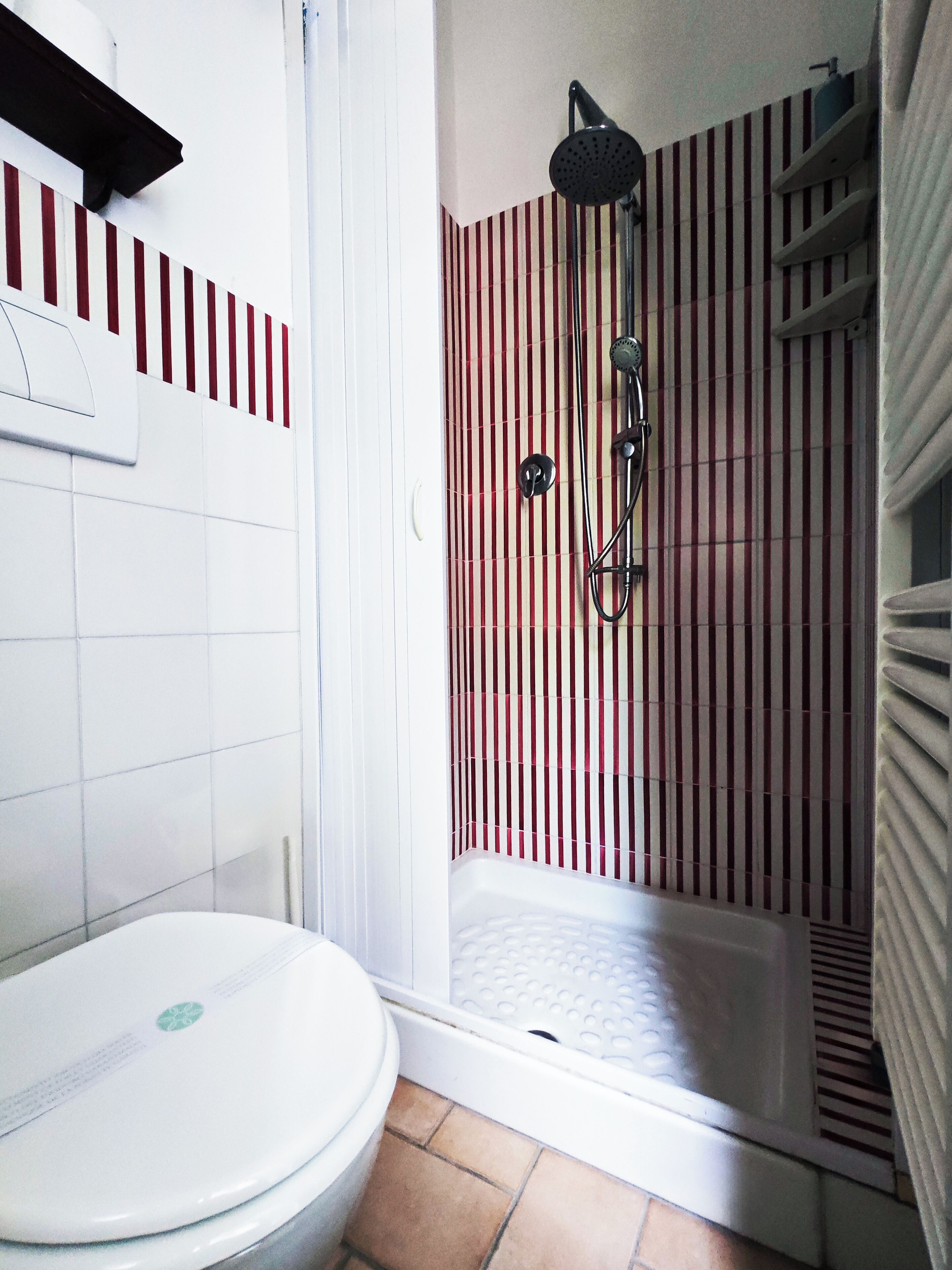 Un baño con inodoro blanco, ducha con pared a rayas rojas y blancas, y bañera blanca.