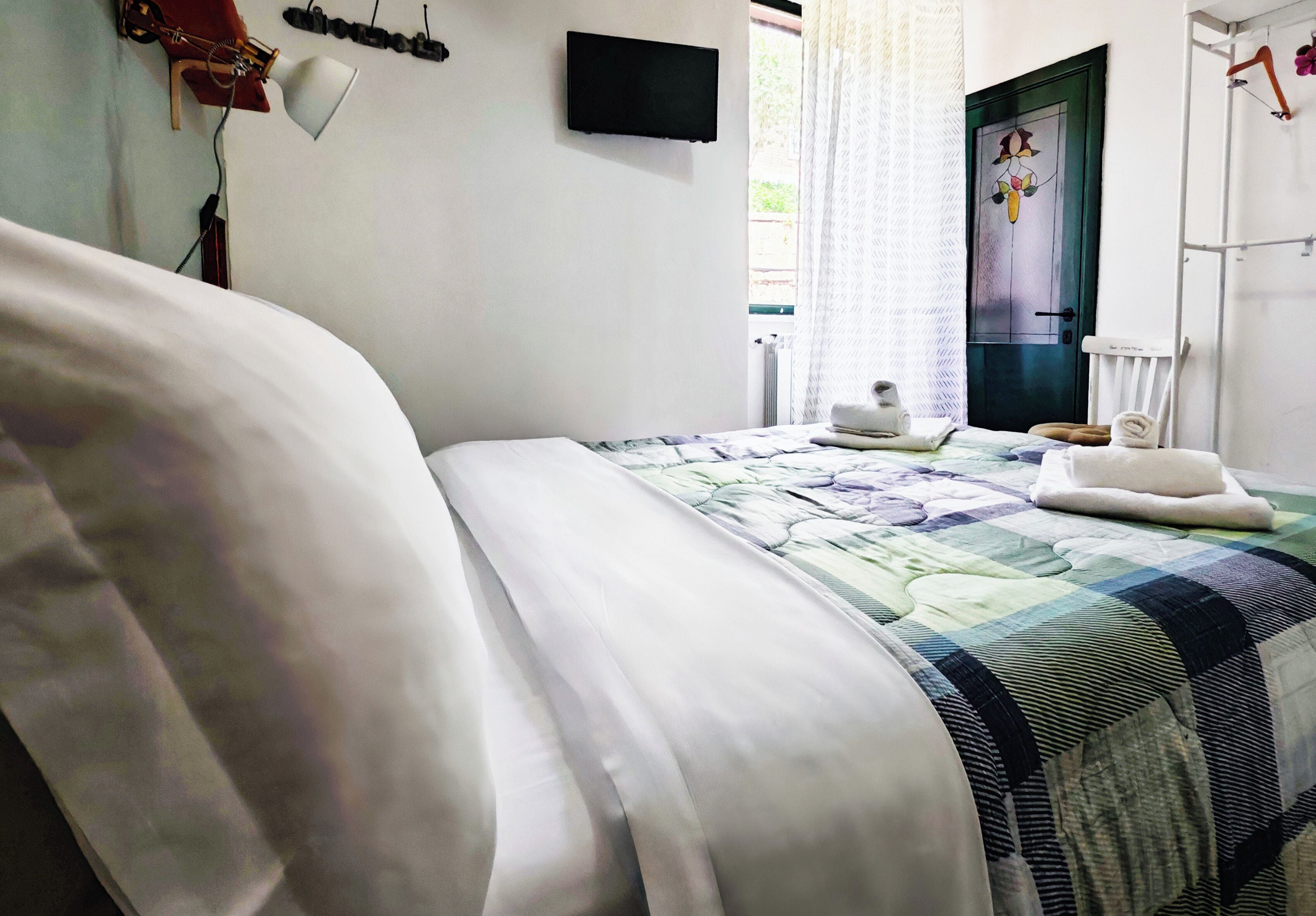A neatly made bed with a quilt, a flat-screen TV mounted on the wall, a window with curtains, and a door with a decorative glass panel.