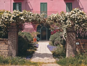 Un camino que lleva a un edificio con fachada rosa, contraventanas verdes y entrada de ladrillo.