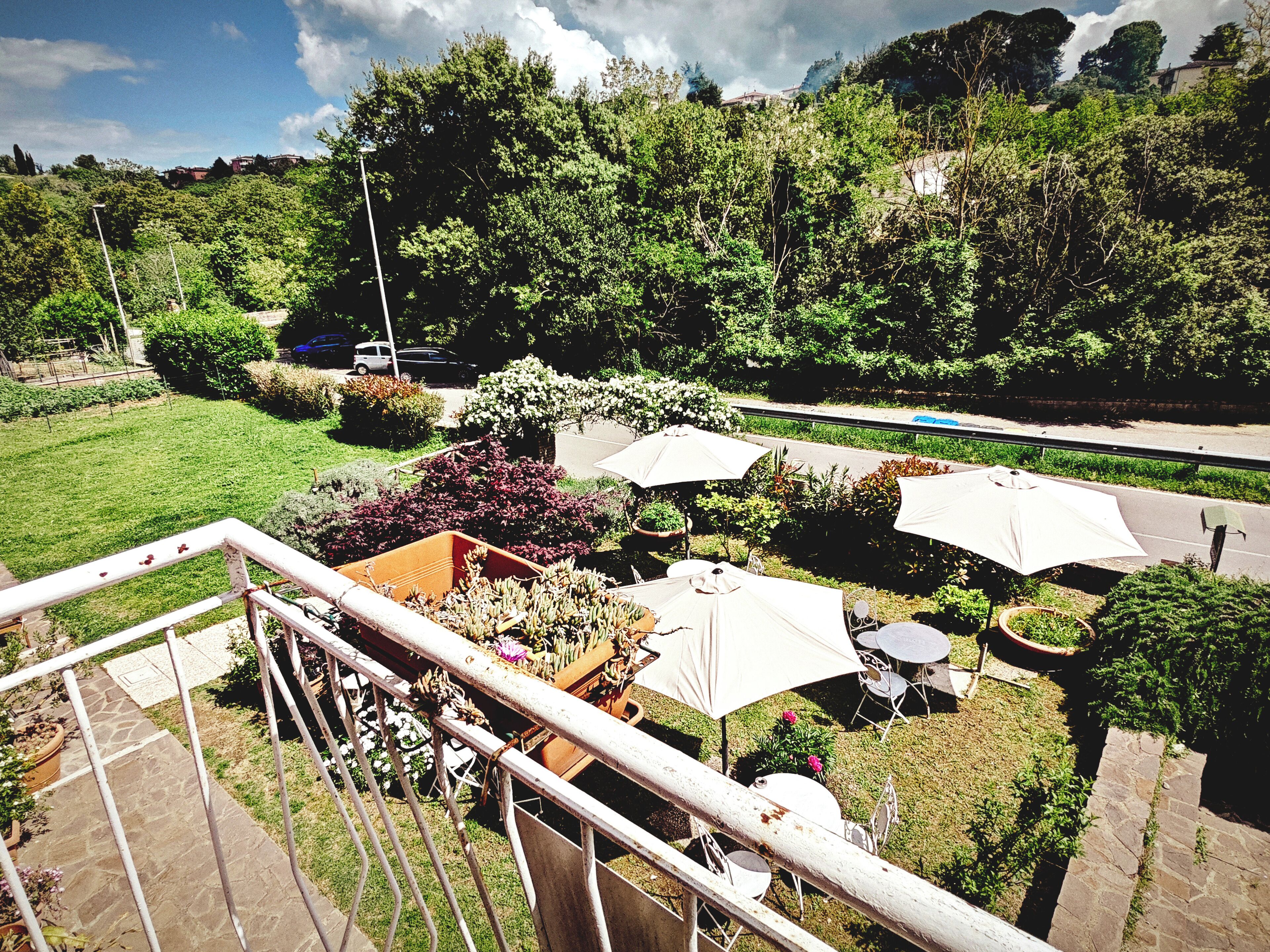 Ein Balkon mit Blick auf einen Garten, eine Sitzgelegenheit im Freien mit Sonnenschirmen und eine Straße im Hintergrund.