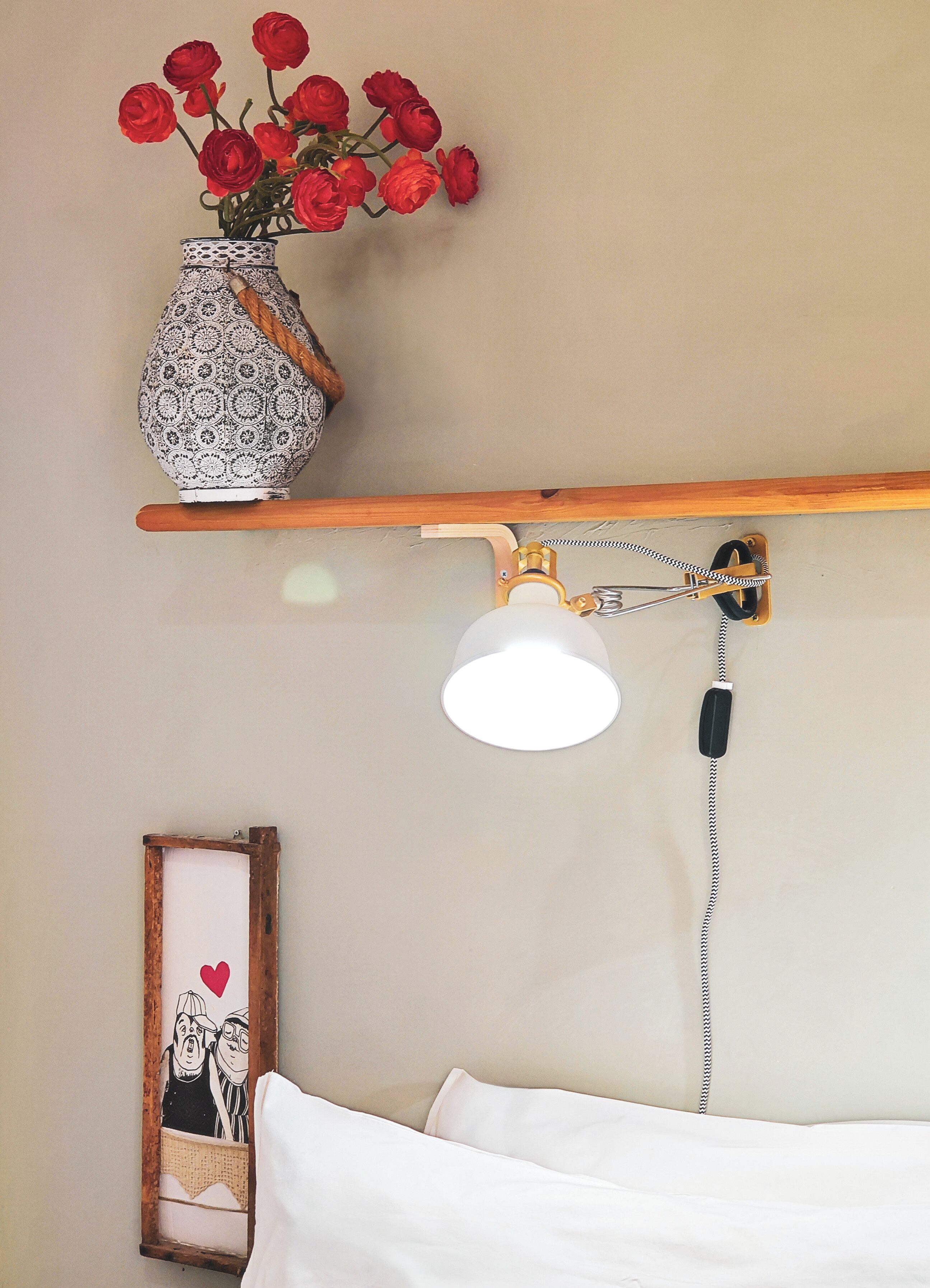 A bed with white bedding, a wall-mounted lamp, a wooden shelf with a vase of red flowers, and a framed picture with a heart.