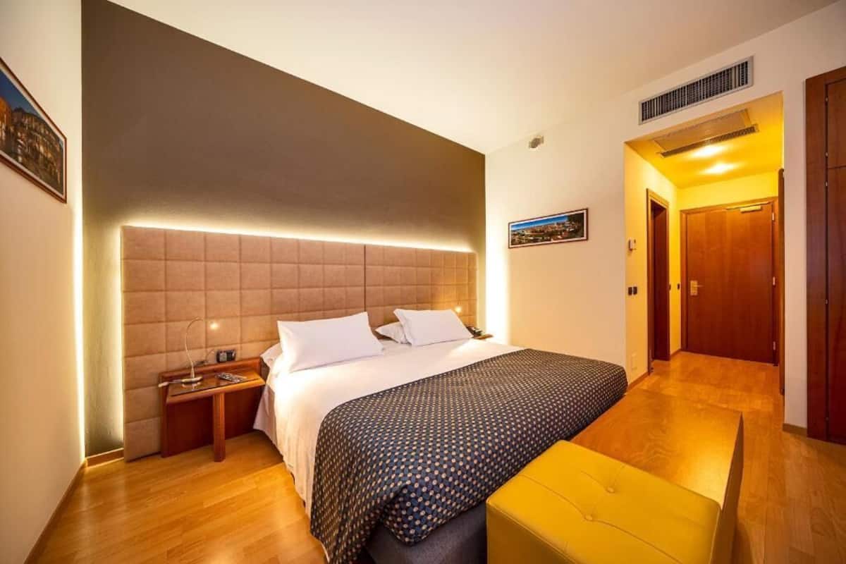 A hotel room with a large bed, a bedside table, a bench, and a door leading to another room.