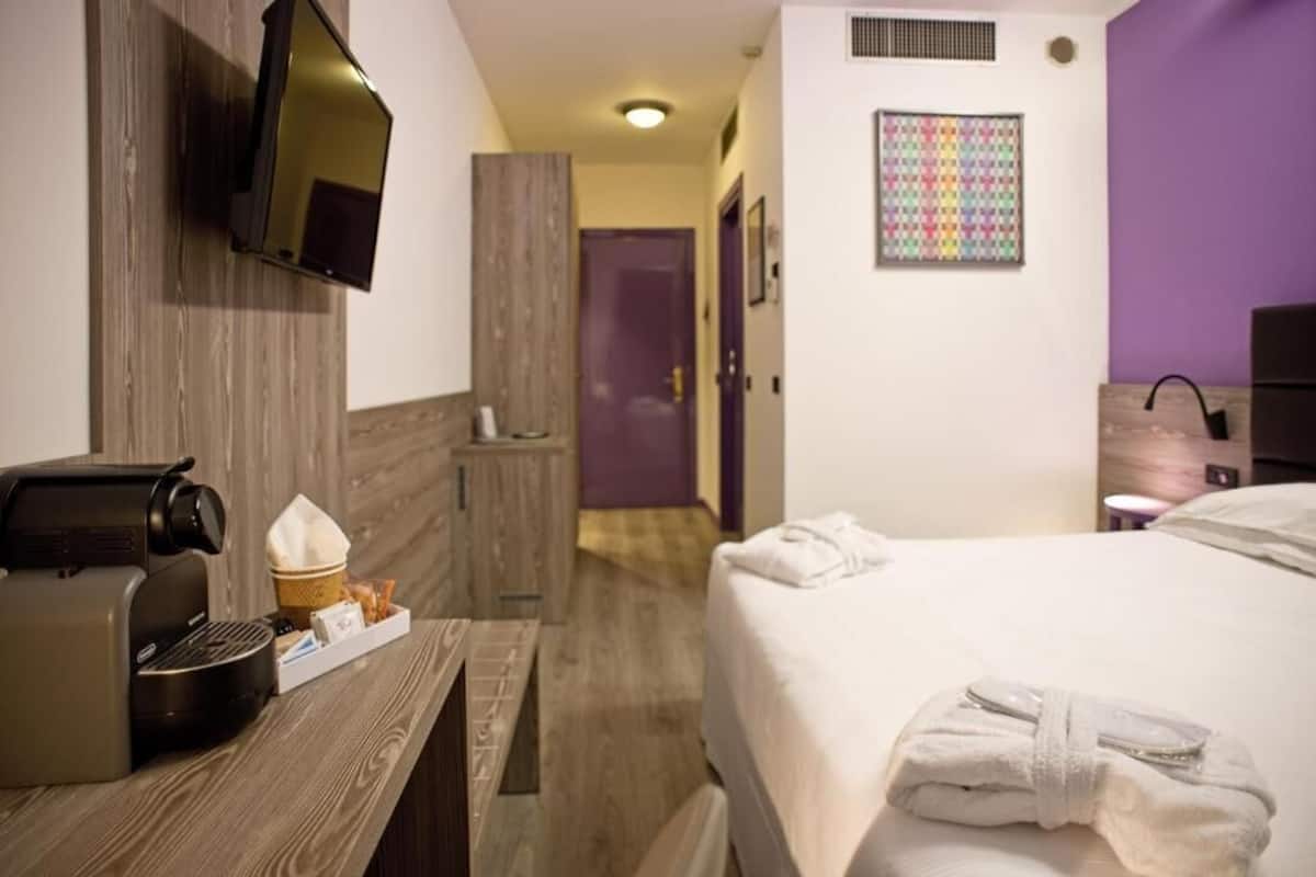 A hotel room with a bed, a coffee machine, a small table with a basket, a TV mounted on the wall, and a colorful artwork on the wall.