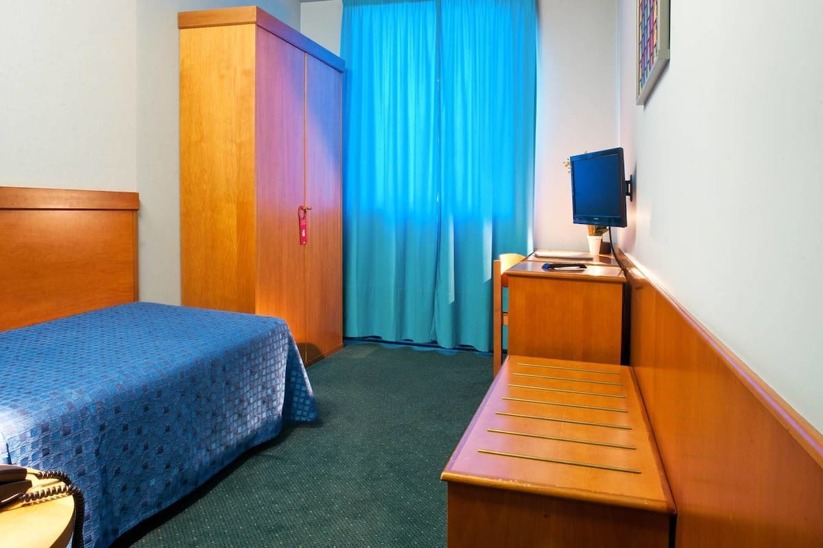 A hotel room with a bed, a wardrobe, a desk with a computer, and a window with curtains.