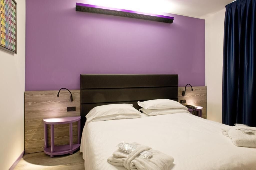 Quarto de hotel com cama, mesa de cabeceira, um banquinho pequeno roxo e luminária fixada na parede.